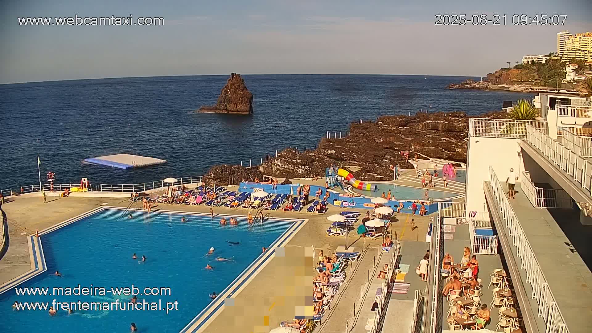 An outdoor swimming pool complex sits on a rocky coastline overlooking a calm ocean under a clear, sunny sky.