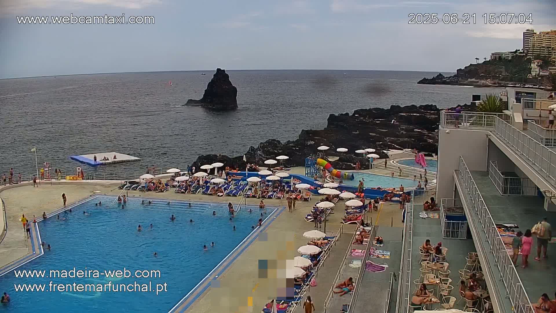 An outdoor pool area with many people swimming and lounging on deck chairs overlooks a rocky coastline and ocean under a mostly sunny sky.