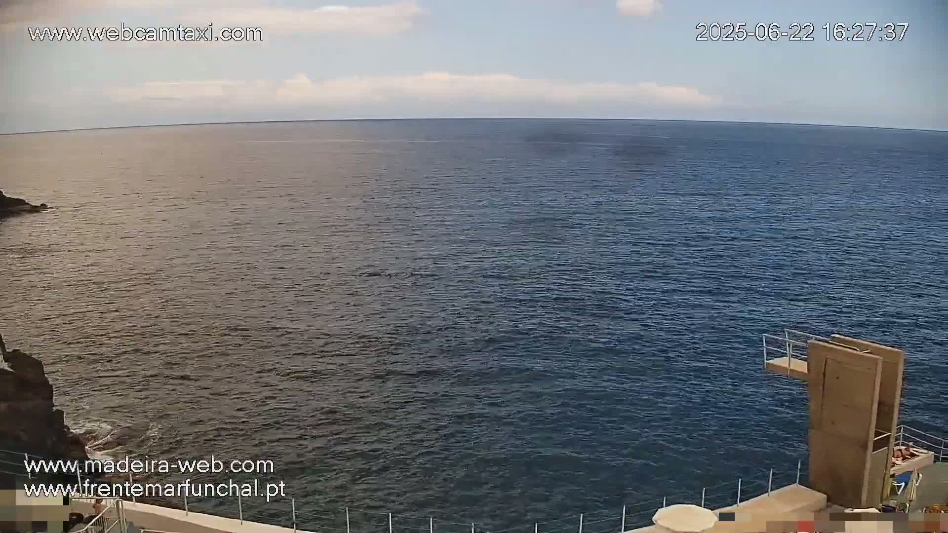 A mostly clear sky overlooks a dark blue ocean, with a diving platform and rocky shoreline visible in the foreground.
