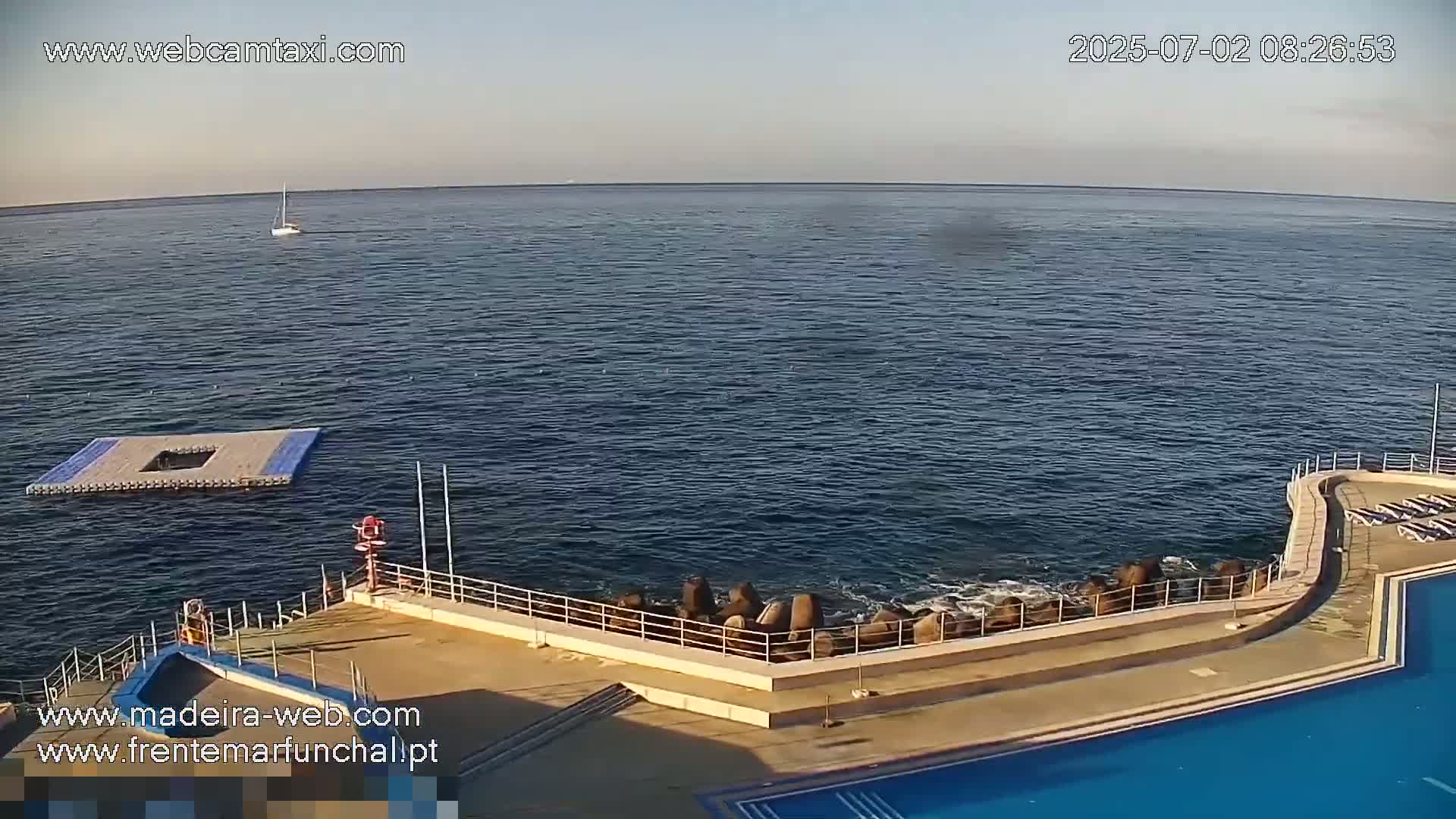 A calm ocean is seen next to a pool area with a railing and breakwater, under a clear sky.
