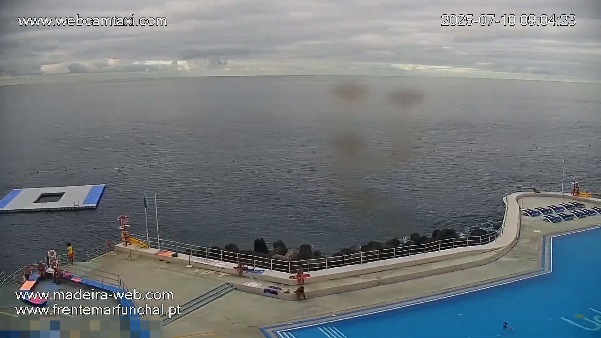 An outdoor pool and a floating platform are situated next to a dark-blue ocean under an overcast sky.
