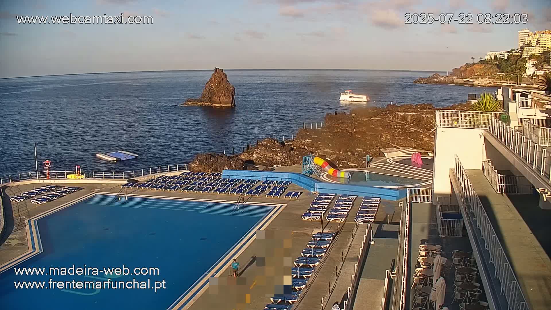 An outdoor pool with surrounding lounge chairs overlooks a rocky coastline and ocean, under a clear sky.