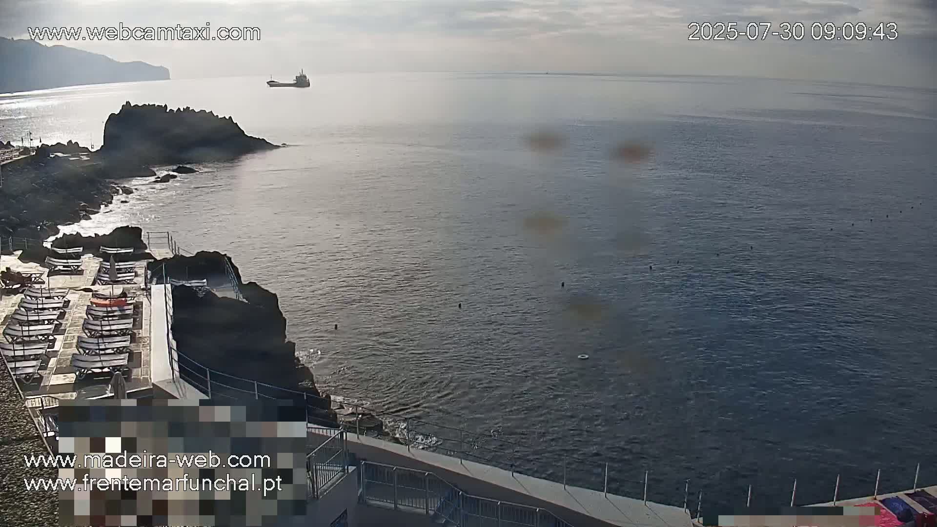 A mostly sunny day shows a rocky coastline with a deck of lounge chairs overlooking a calm ocean with a distant ship.
