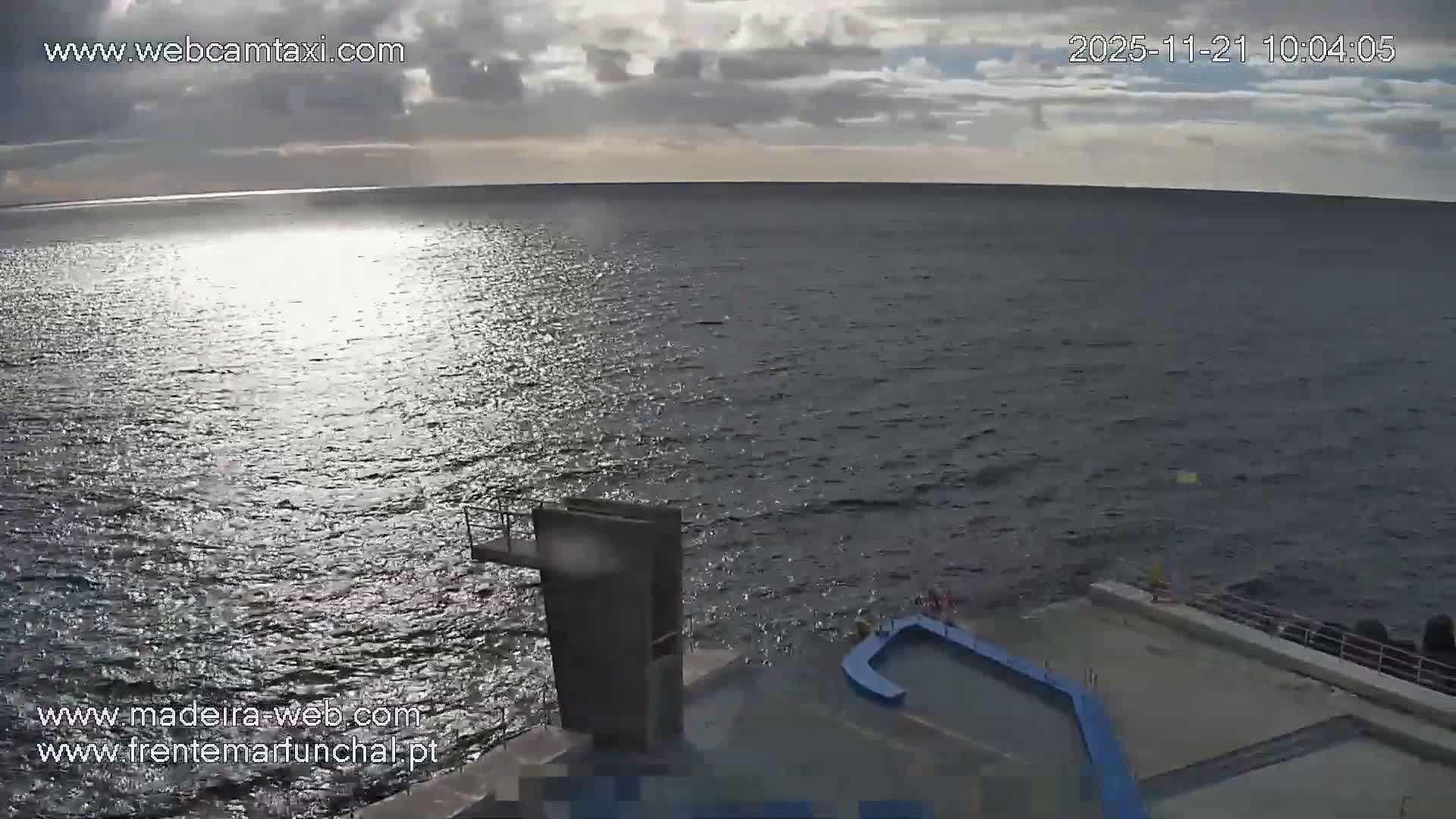 A partly cloudy day with bright sun reflecting across a vast expanse of dark blue water, featuring a concrete diving platform and a curved blue structure in the foreground.