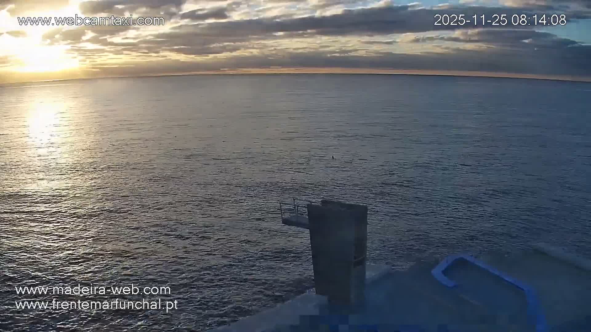 A hazy sunrise illuminates a calm, rippled ocean under a partly cloudy sky, with a concrete diving board structure extending over the water and a blue handrail visible in the foreground.