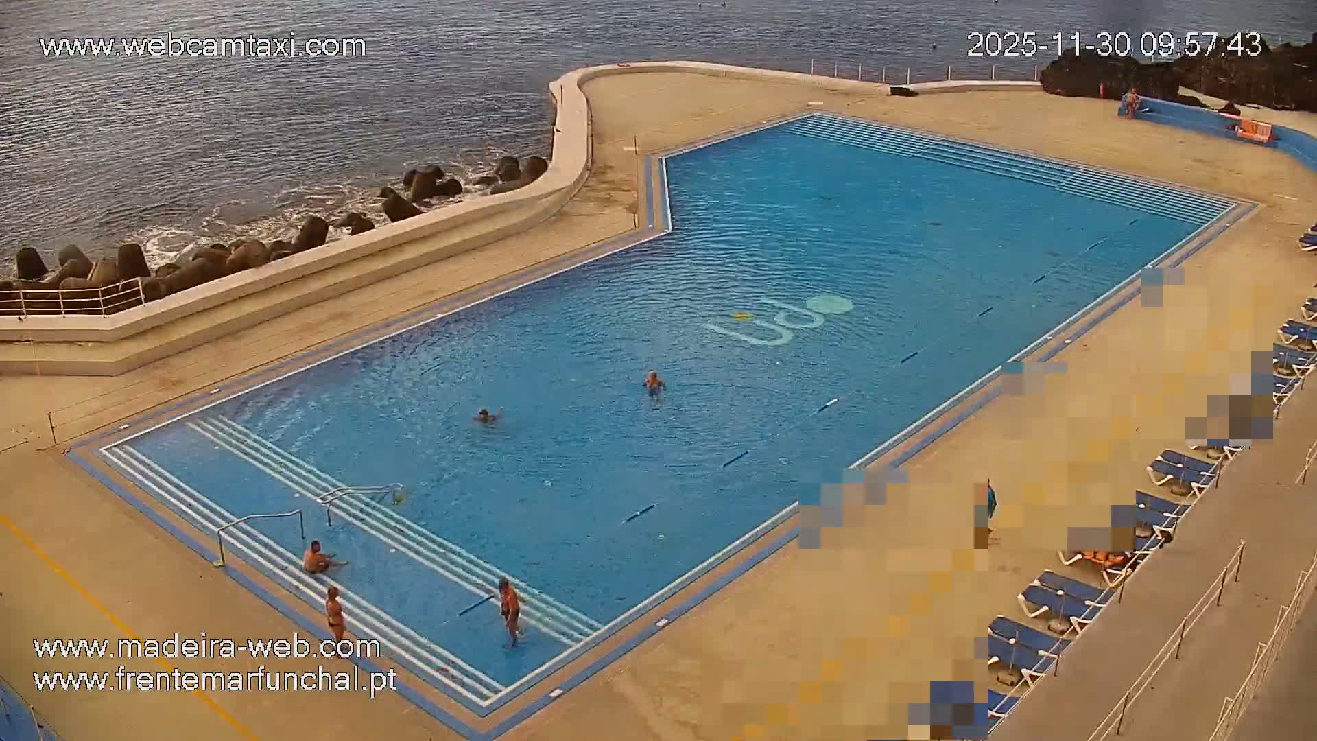 Several people are seen swimming and relaxing around a large outdoor swimming pool complex situated beside the ocean, which features a rock barrier and gentle waves, under clear daytime conditions.