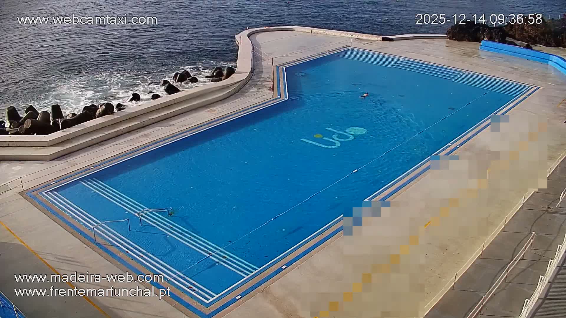 Several people are seen swimming and relaxing around a large outdoor swimming pool complex situated beside the ocean, which features a rock barrier and gentle waves, under clear daytime conditions.