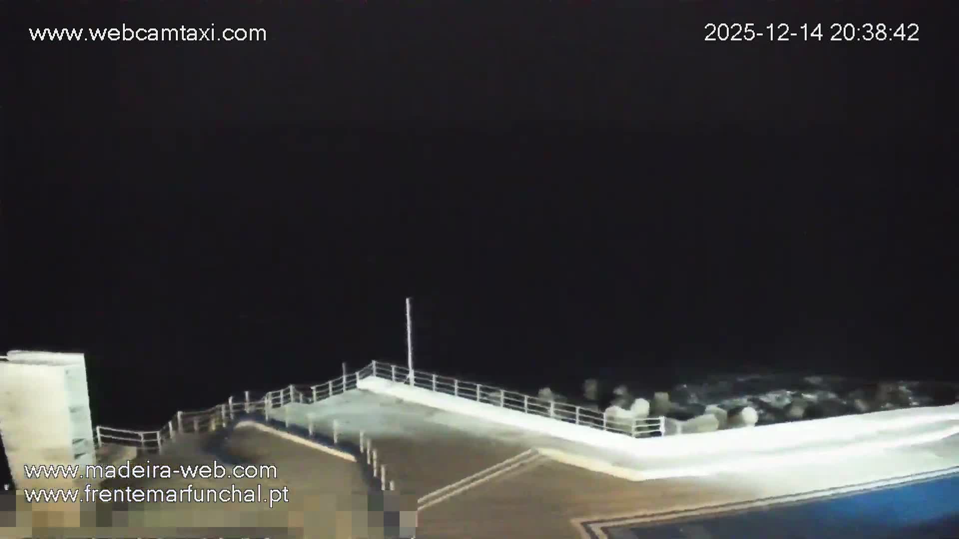 Several people are seen swimming and relaxing around a large outdoor swimming pool complex situated beside the ocean, which features a rock barrier and gentle waves, under clear daytime conditions.
