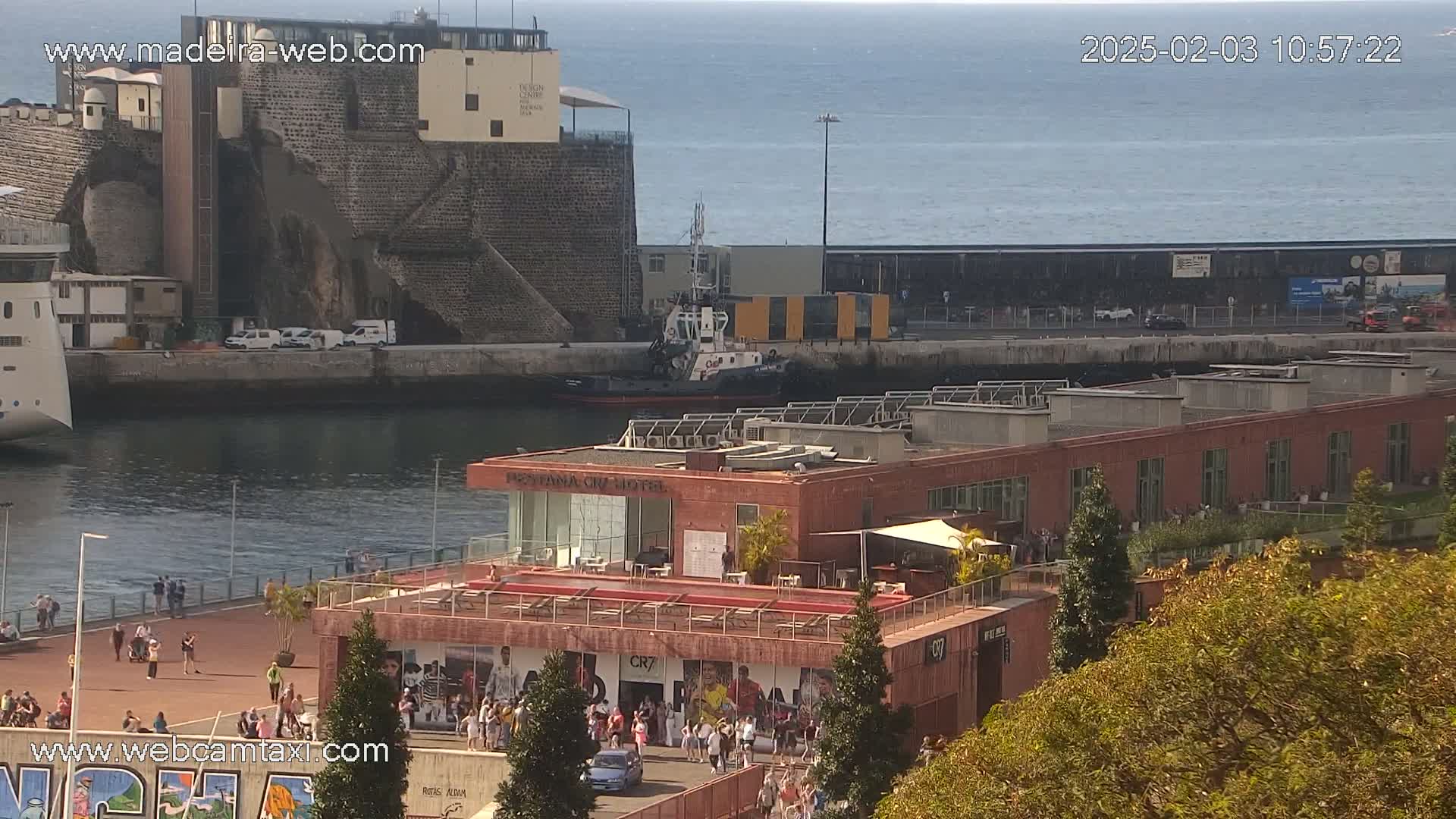 An aerial view shows a harbor scene on a sunny day, with a tugboat in the water near a building with a rooftop patio and people gathered on a promenade.