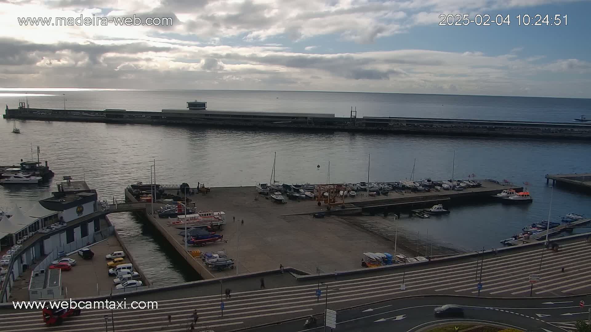 An overcast day shows a harbor filled with numerous boats and yachts, alongside a waterfront road and buildings.