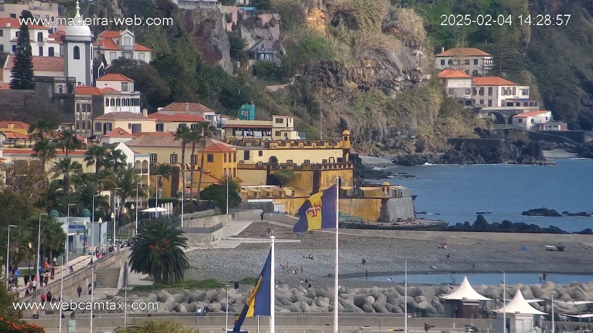 A coastal town with colorful buildings on a hillside overlooking a pebble beach and calm ocean under a clear sky.