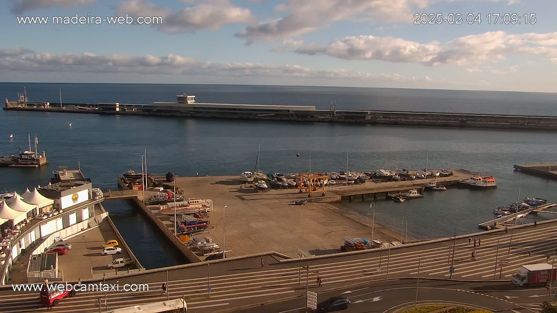 An aerial view of a harbor filled with boats on a partly cloudy day, overlooking a calm ocean.