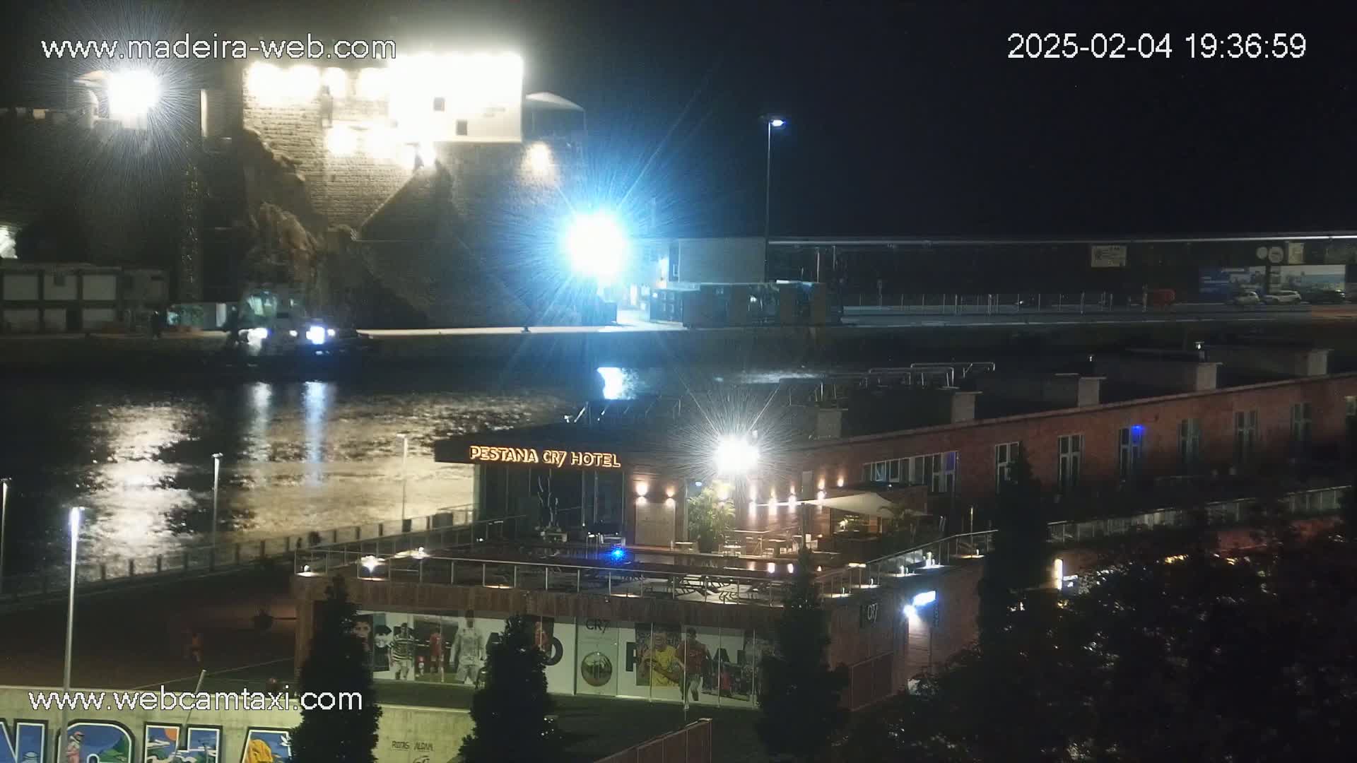 A nighttime view of a waterfront scene shows a calm body of water reflecting lights, a stone building on a hill, a hotel with outdoor seating, and a roadway above.