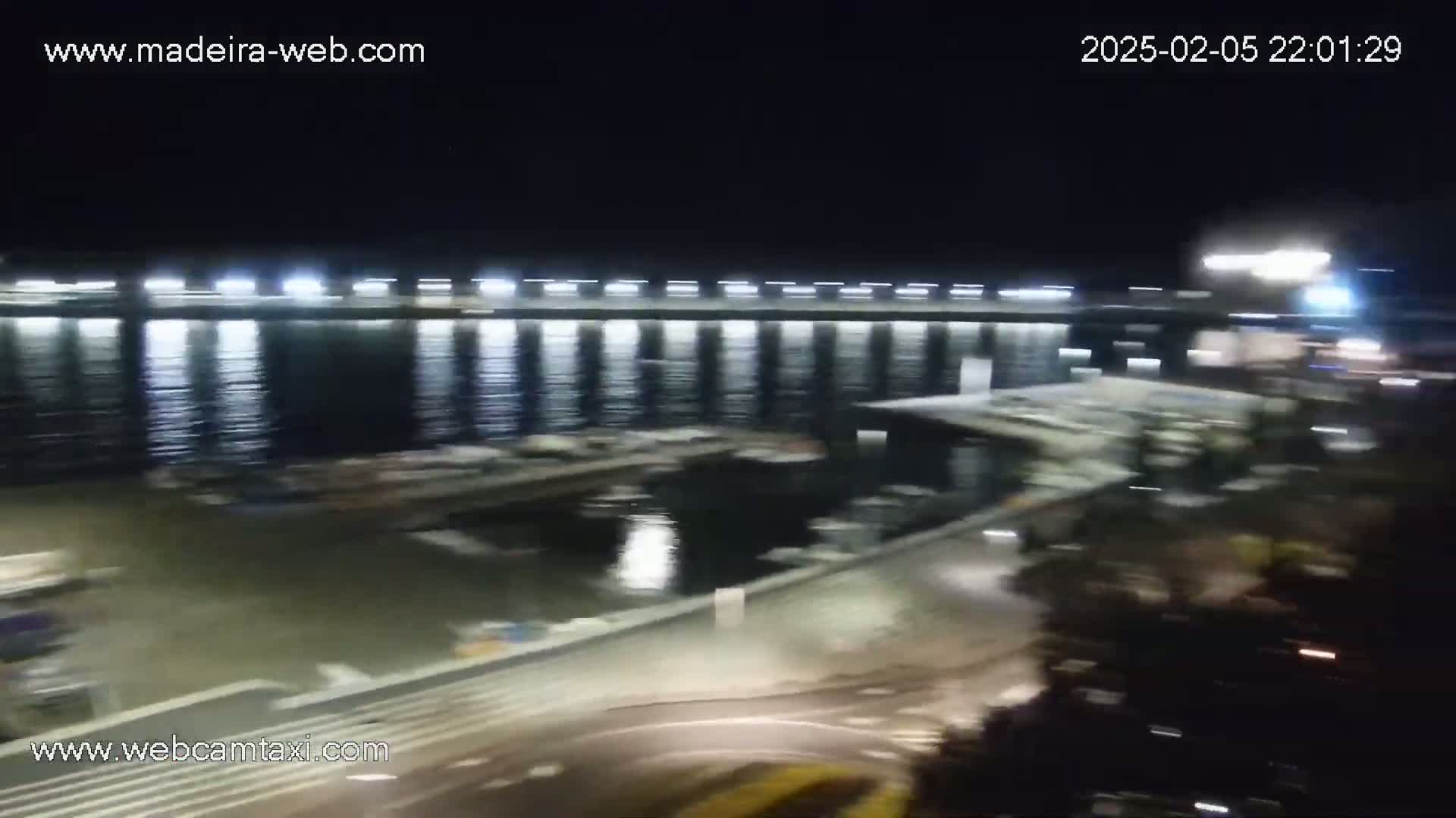 A nighttime view of a marina with boats docked under a brightly lit pier, and calm water reflecting the lights.