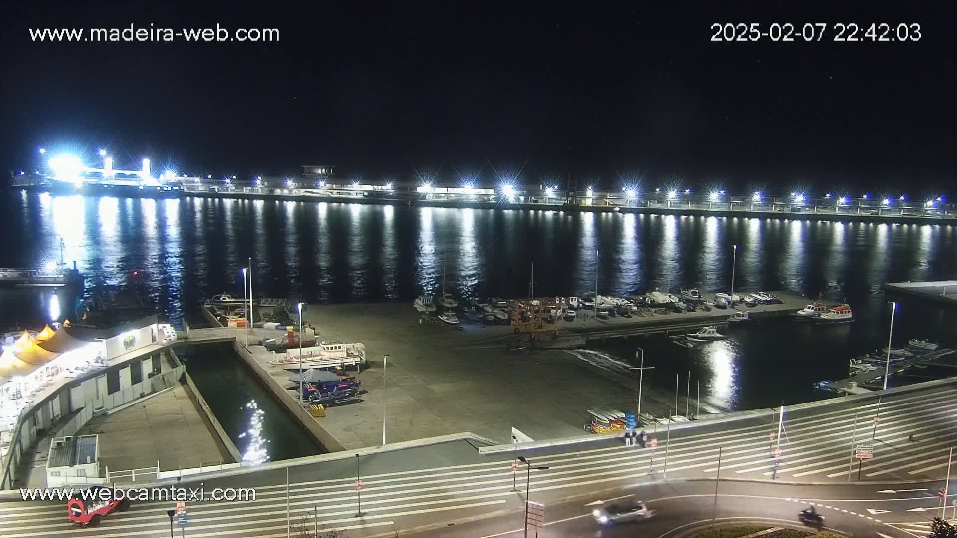 A nighttime view of a harbor filled with boats, lit by bright lights reflecting on the calm water.