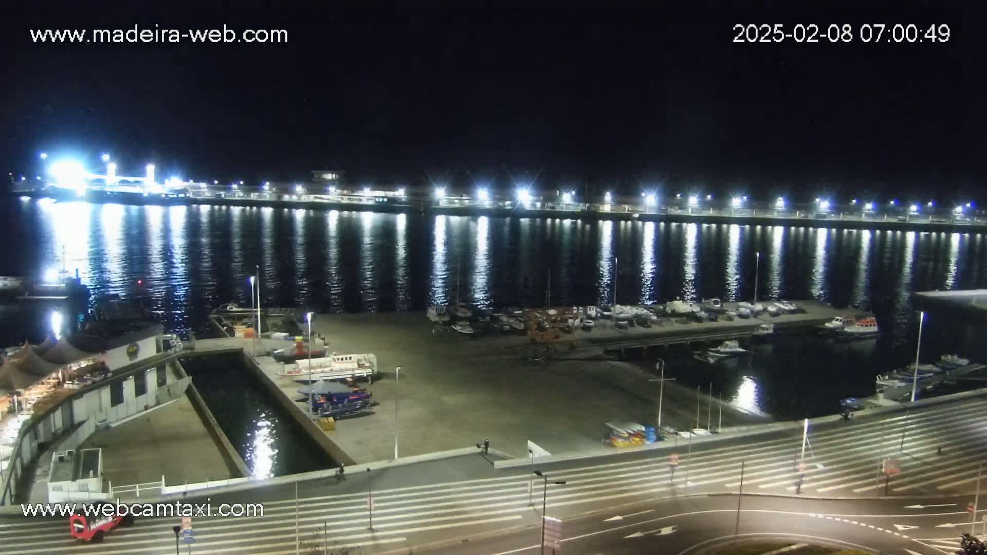 A brightly lit harbor at night shows numerous boats docked along a pier, with calm water reflecting the lights.