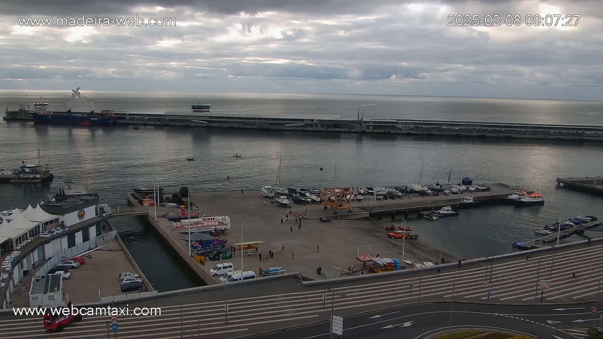 An overcast day reveals a harbor filled with numerous boats and ships, alongside a dock with people and vehicles.