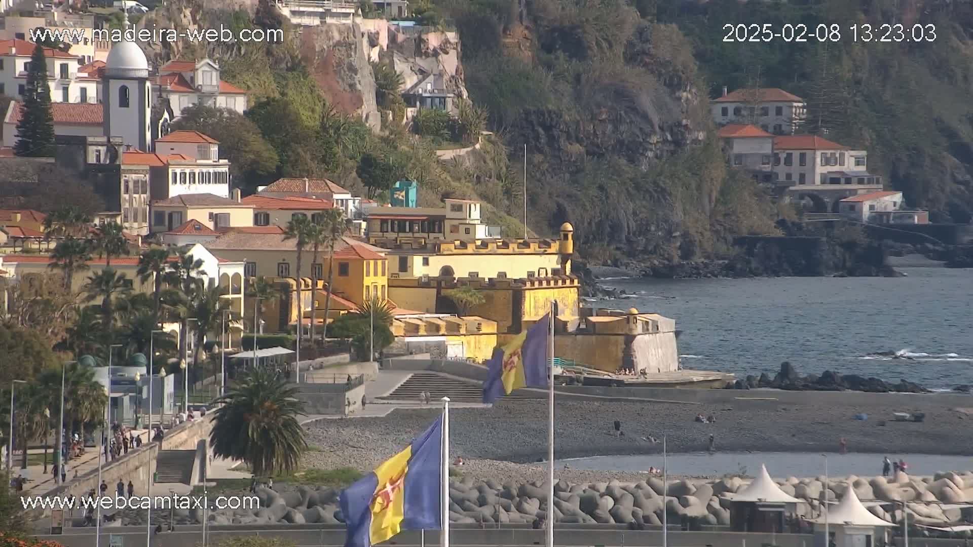 A seaside town with colorful buildings, a stone fortress, and a dark-sand beach on a sunny day.