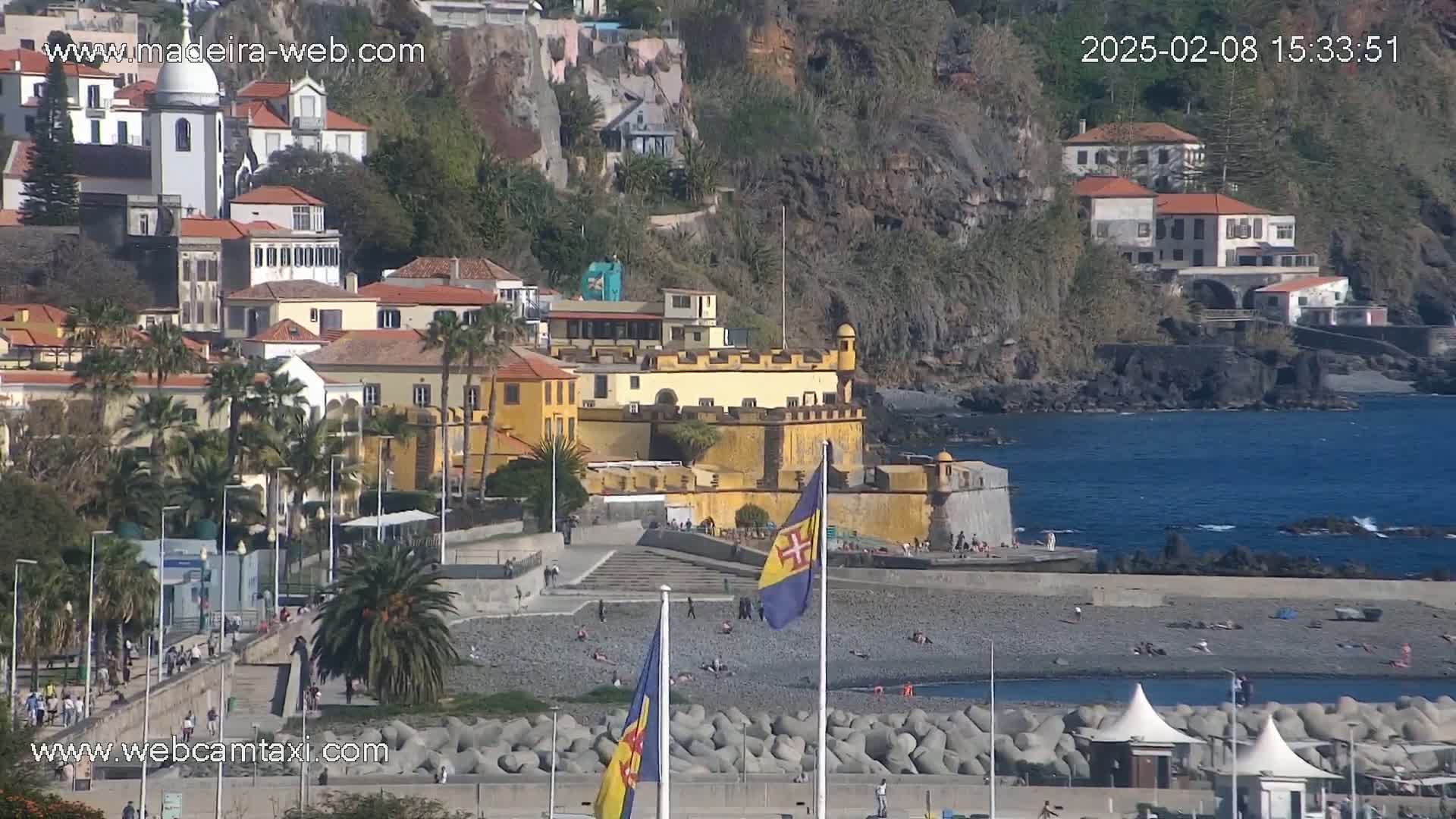 A coastal town with yellow buildings, a fortress, and a pebble beach is seen on a sunny day.