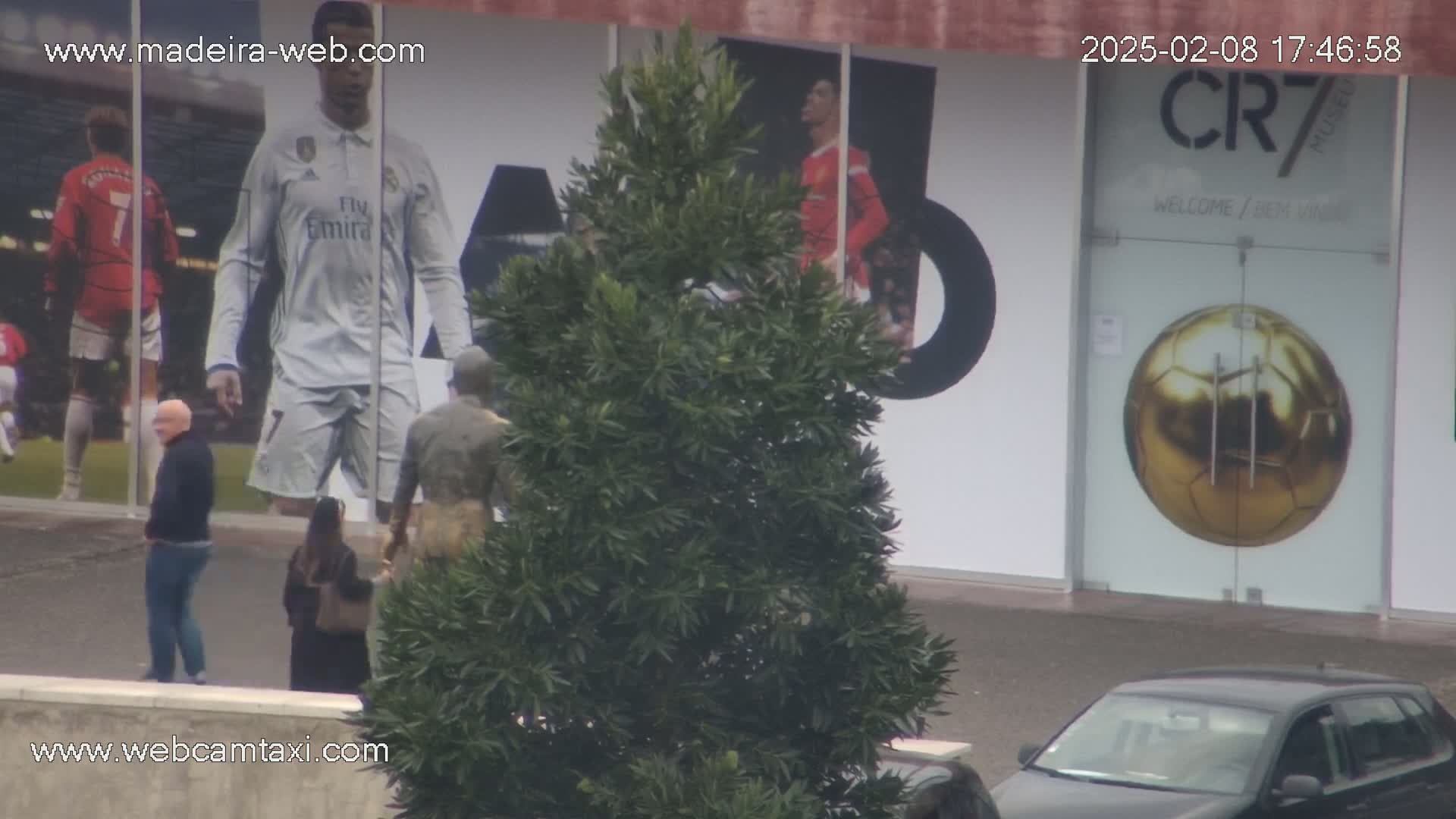 A large building with images of soccer players on the exterior, a man and woman walking in front, a small tree partially obscuring the view, and a car parked in the foreground on an overcast day.