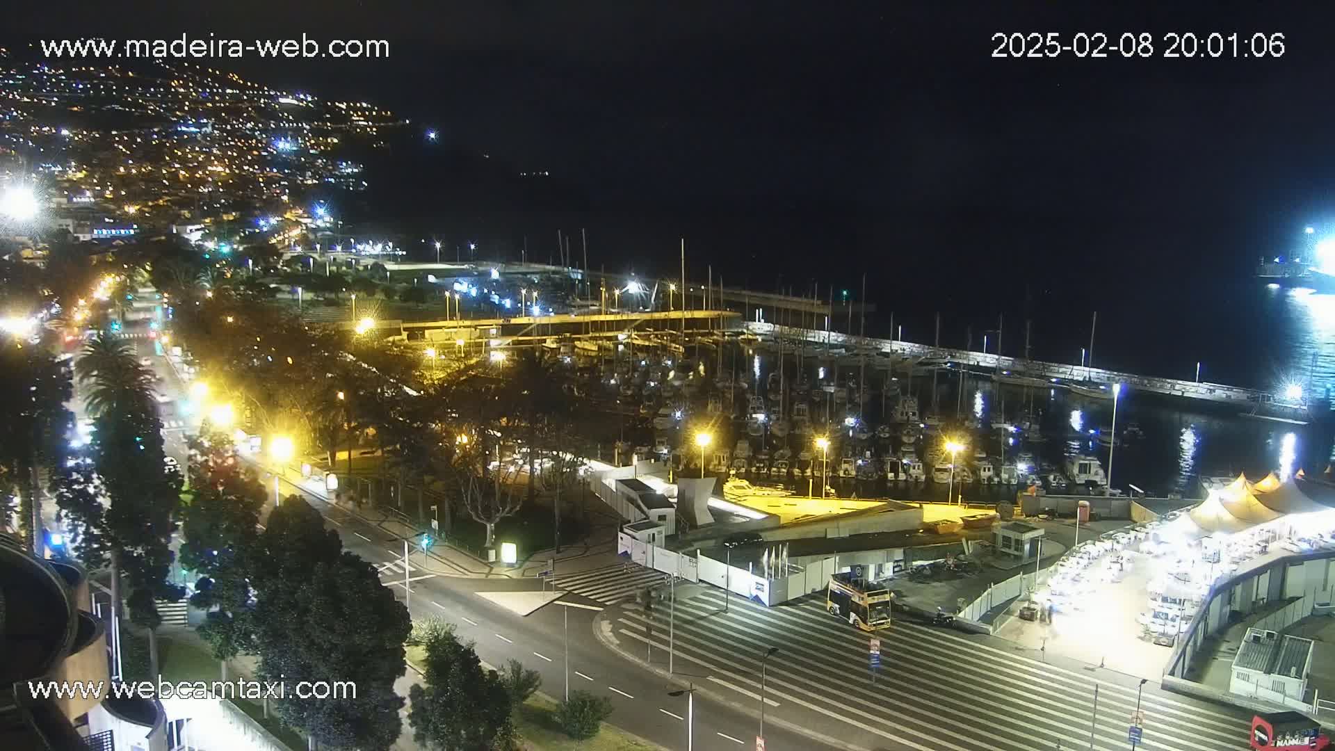 A nighttime aerial view of a harbor filled with boats, adjacent to a city with illuminated streets and buildings, under a clear, dark sky.
