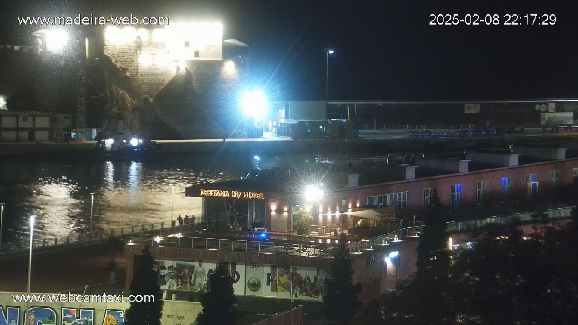 A nighttime view of a waterfront area shows a hotel, a body of water reflecting lights, and a stone structure in the distance.