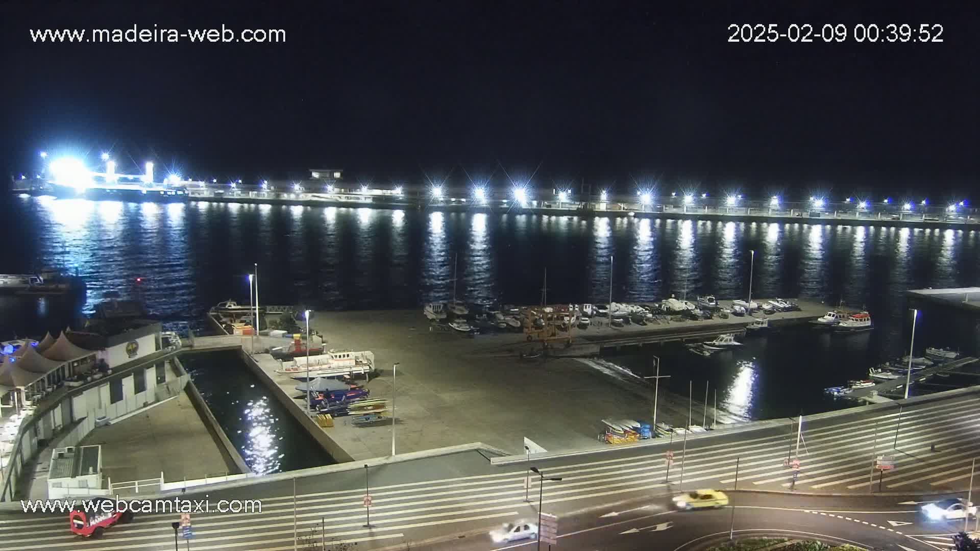 A brightly lit marina at night, with calm water reflecting the lights, and a few cars driving on a road nearby.