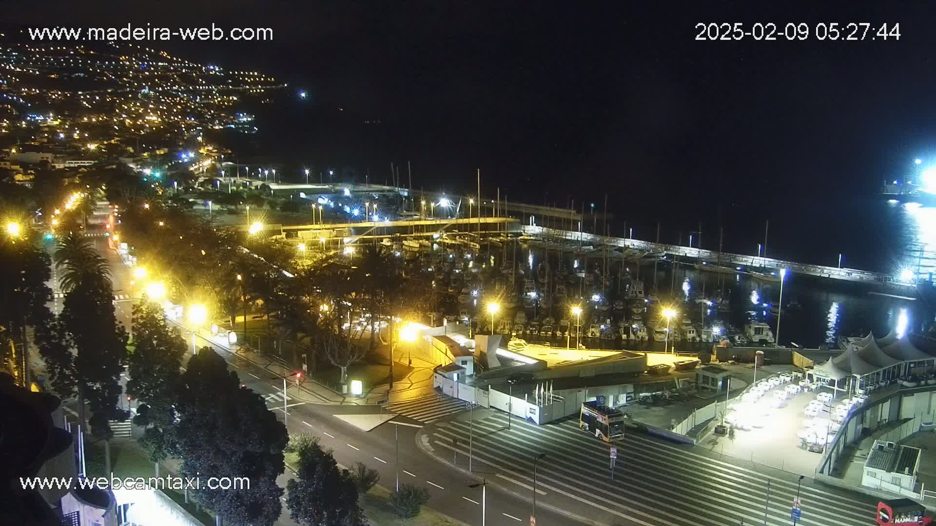 A nighttime, aerial view of a harbor filled with boats, a roadway with cars, and a city illuminated in the background.