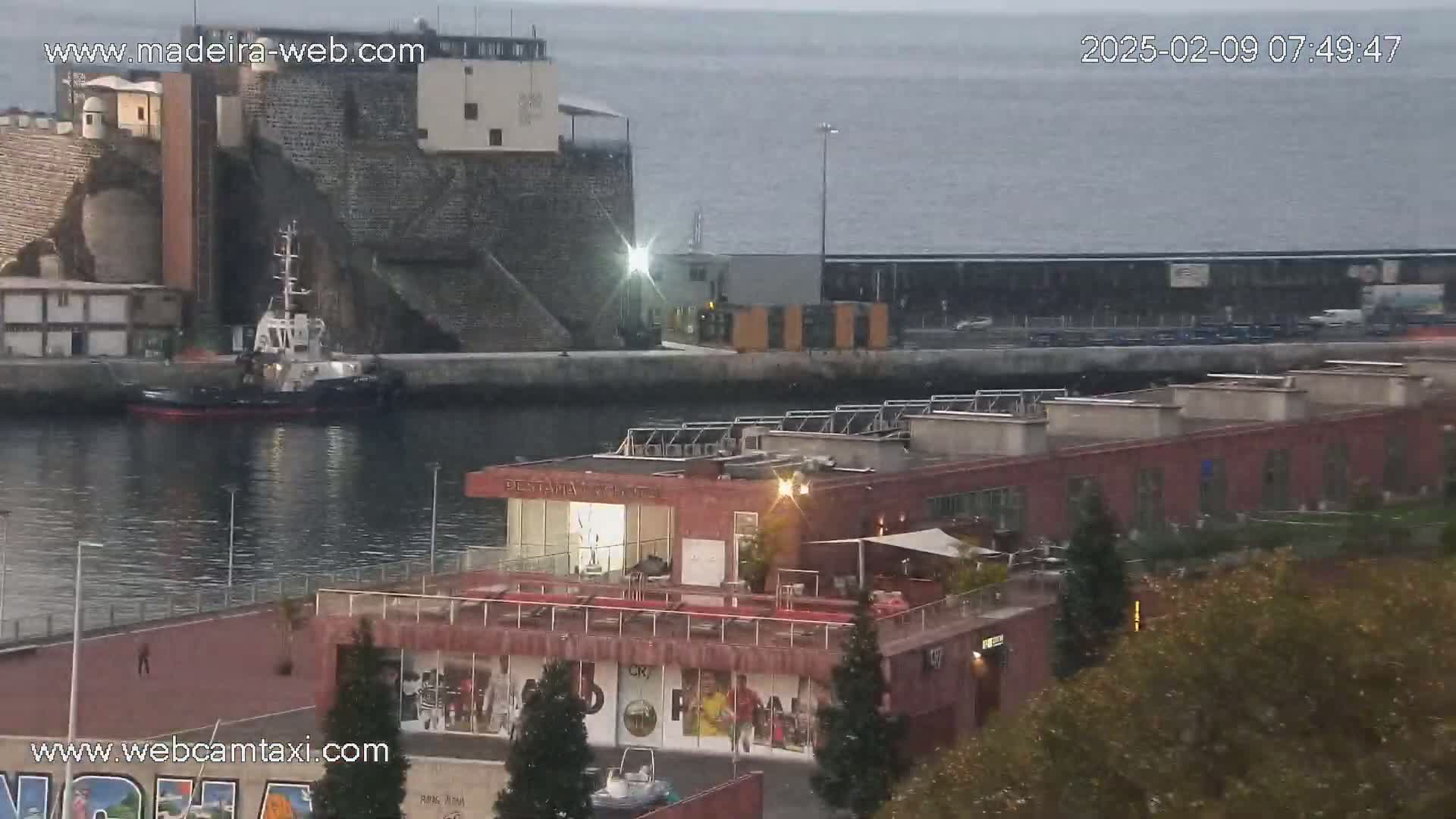 A harbor scene on an overcast day shows a tugboat in the water near a stone building and a red building with a rooftop patio, surrounded by trees and other structures.
