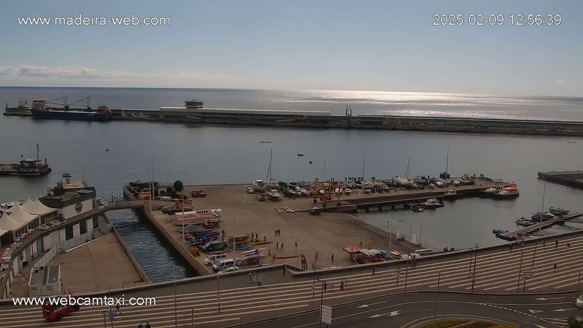 An aerial view of a harbor filled with various boats and ships on a sunny day, with a calm ocean extending to the horizon.