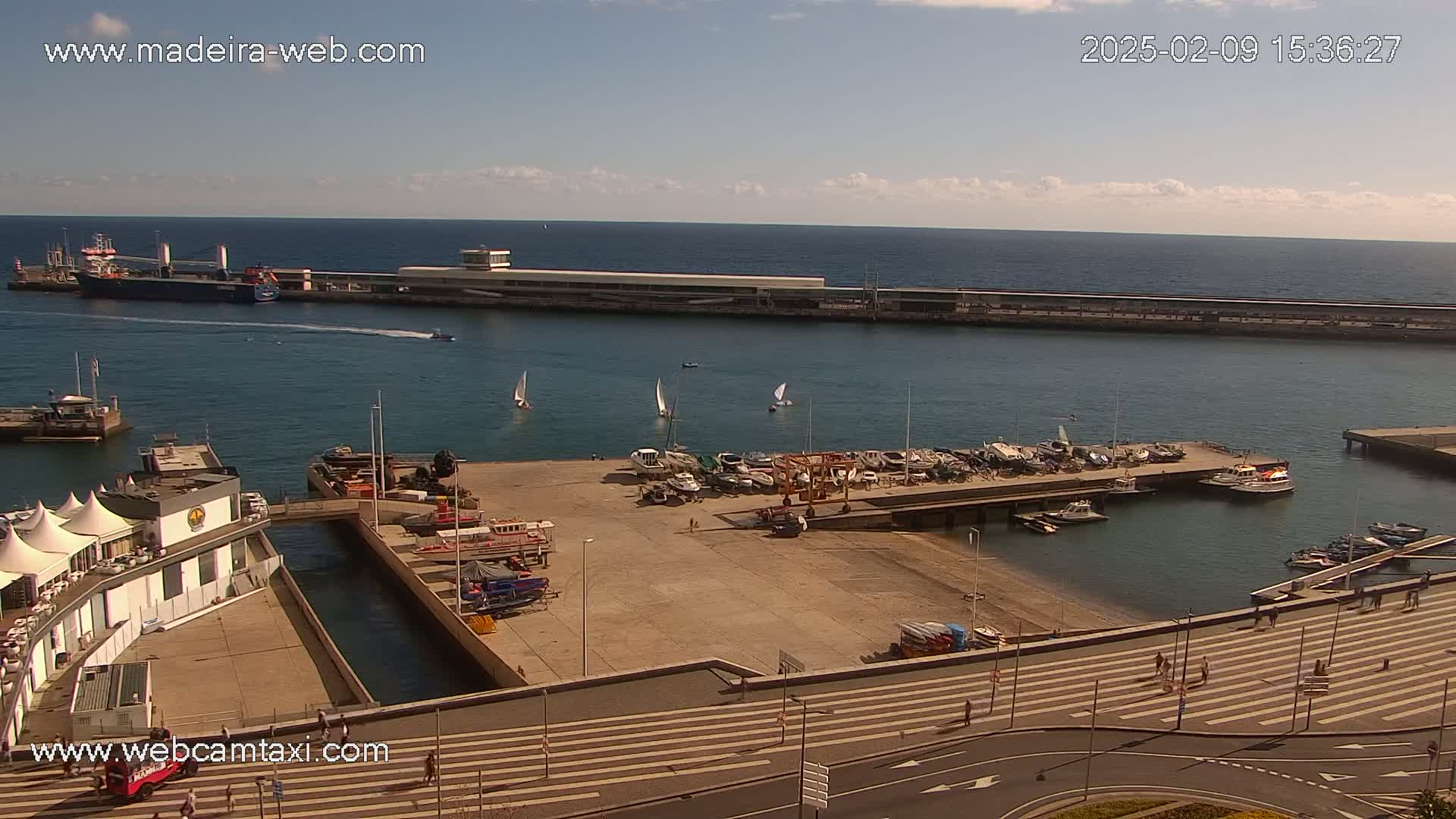 A harbor scene on a sunny day shows a large cargo ship, several sailboats, numerous smaller motorboats docked, and a paved area with pedestrians.