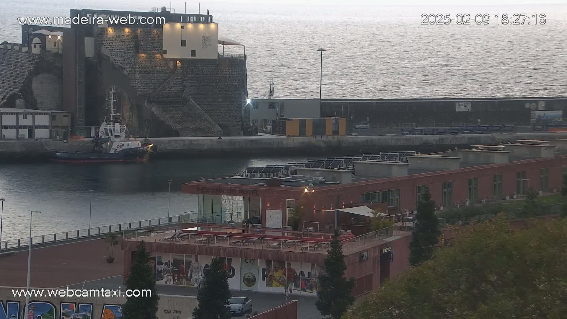 A tugboat is docked near a stone structure and a modern hotel, overlooking calm water under a dusky sky.
