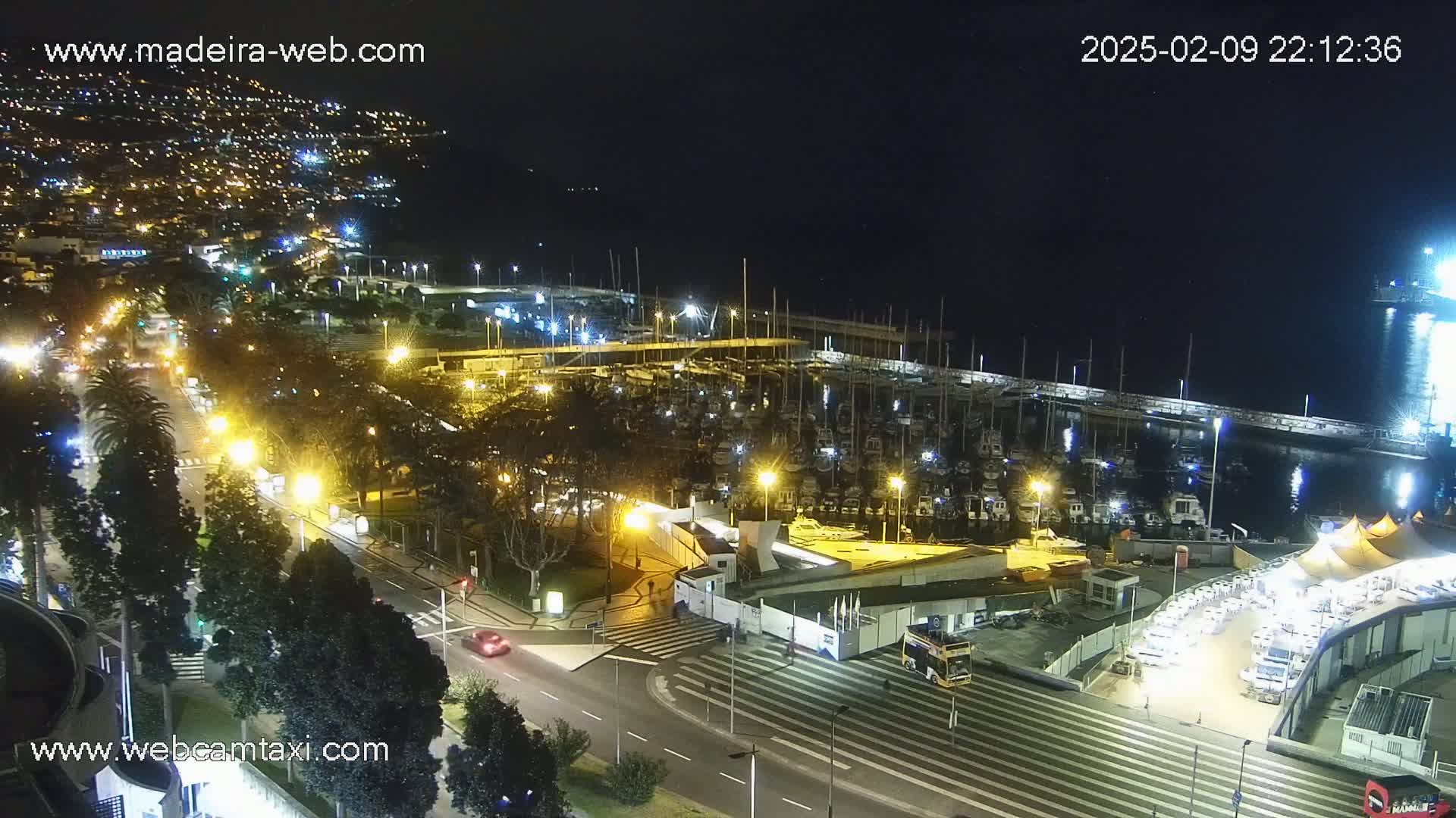 A nighttime aerial view of a harbor filled with boats, adjacent to a city with illuminated streets and buildings under a clear, dark sky.