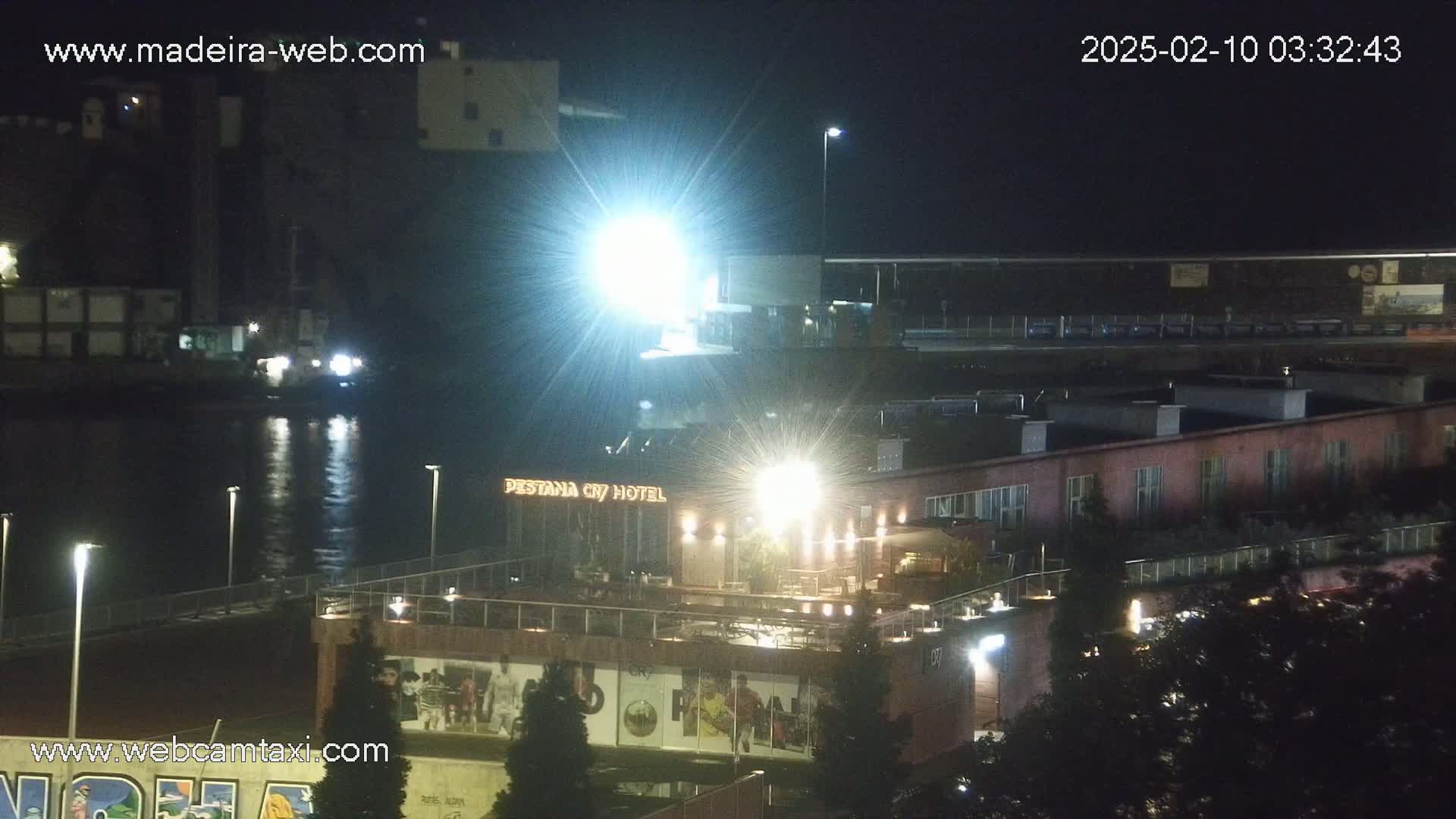A brightly lit waterfront hotel at night, with a body of water reflecting lights and a large building in the background under a dark sky.