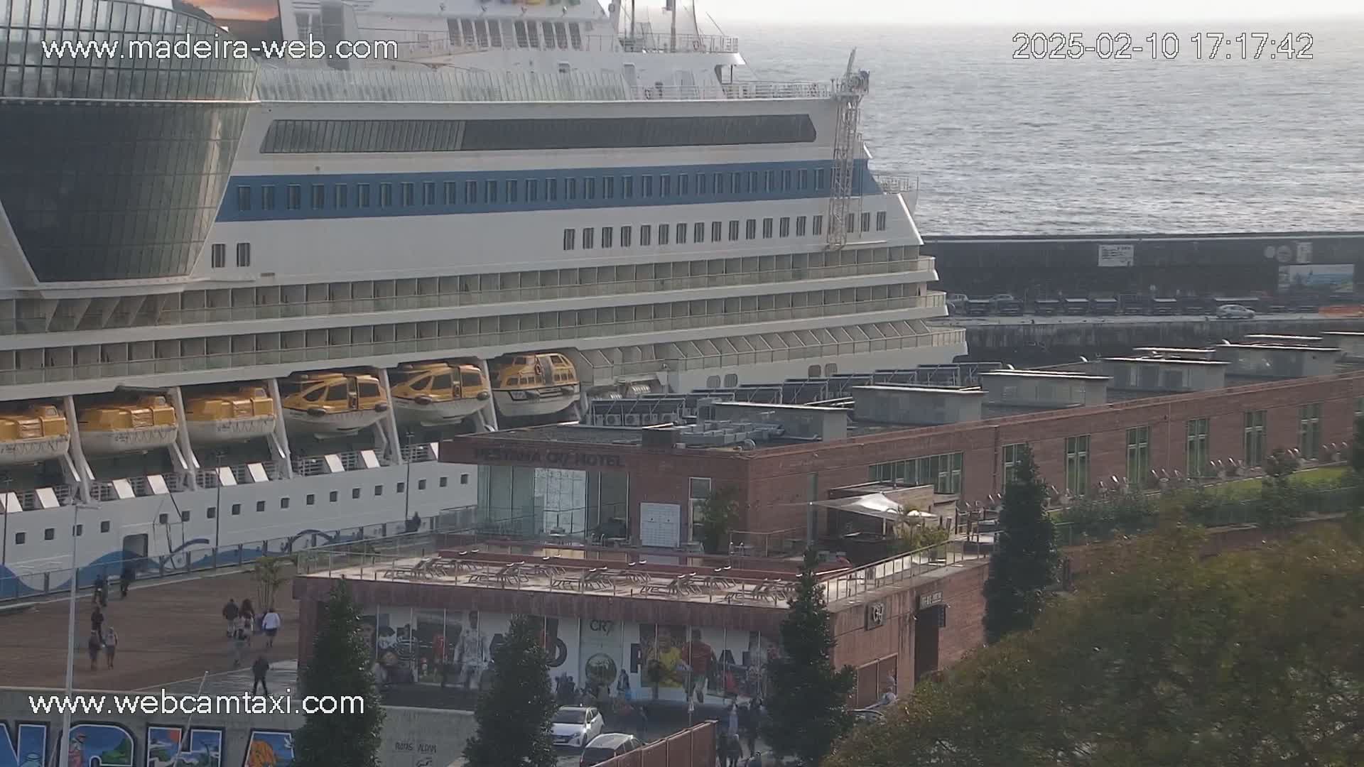 A large cruise ship is docked at a port on a somewhat overcast day, with lifeboats visible on its side, and a brick building with a rooftop terrace is adjacent to the dock.