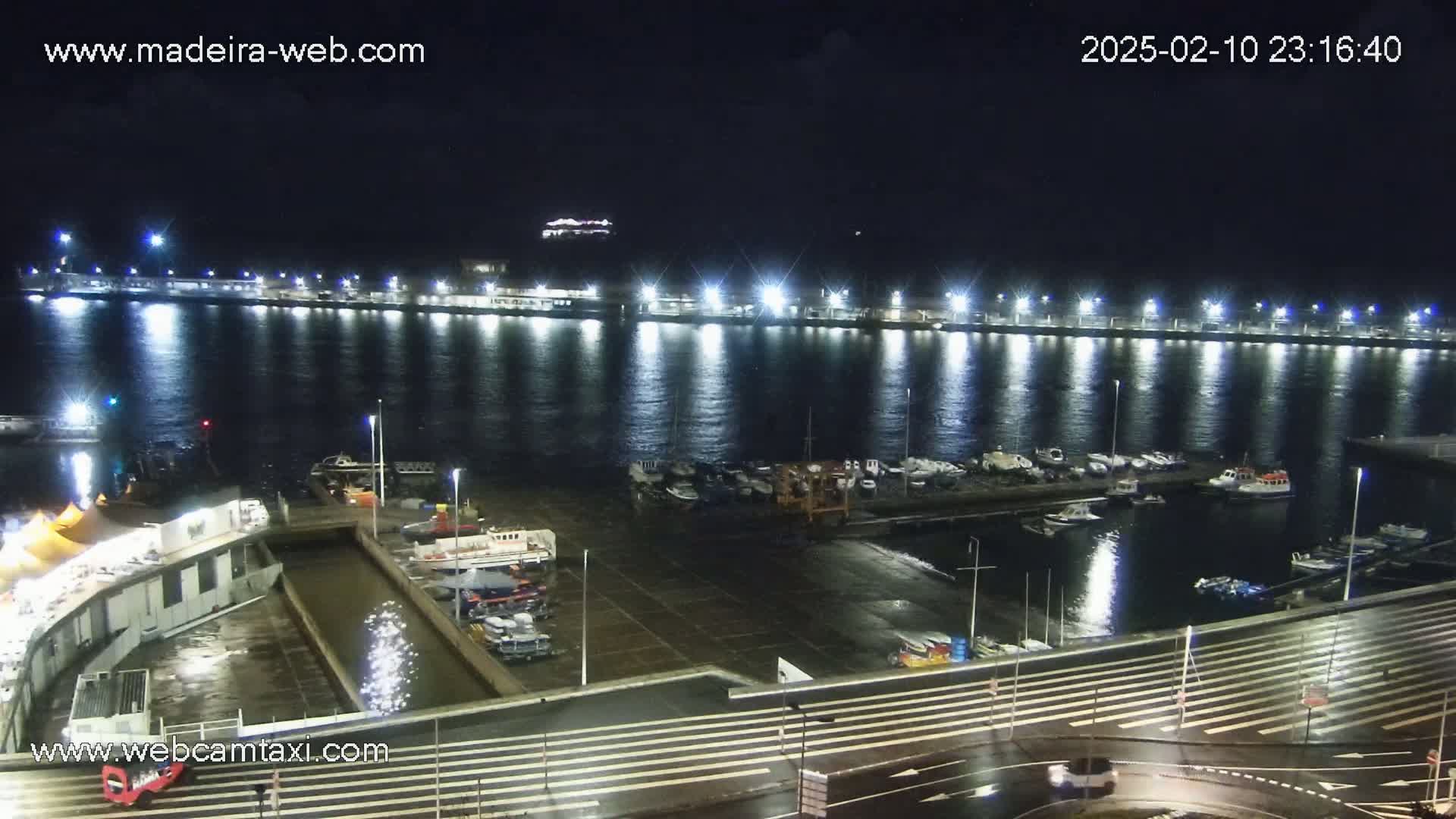 A nighttime view of a harbor filled with boats, illuminated by bright lights reflecting on the calm water.