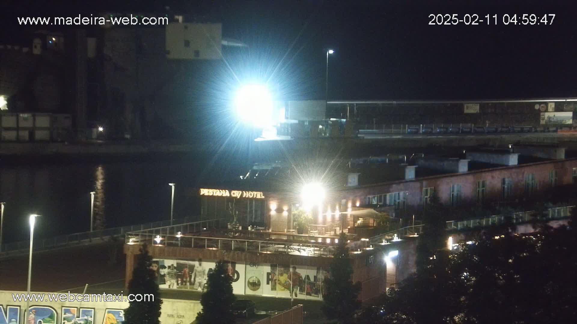 A nighttime view of a waterfront area with brightly lit buildings, including a hotel, and a dark body of water reflecting light.