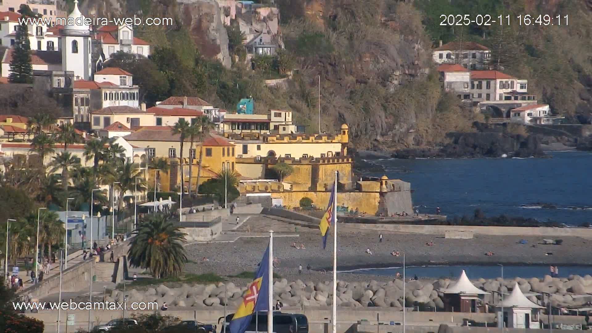 A coastal town with a yellow fortress, palm trees, and a pebble beach, on a sunny day.