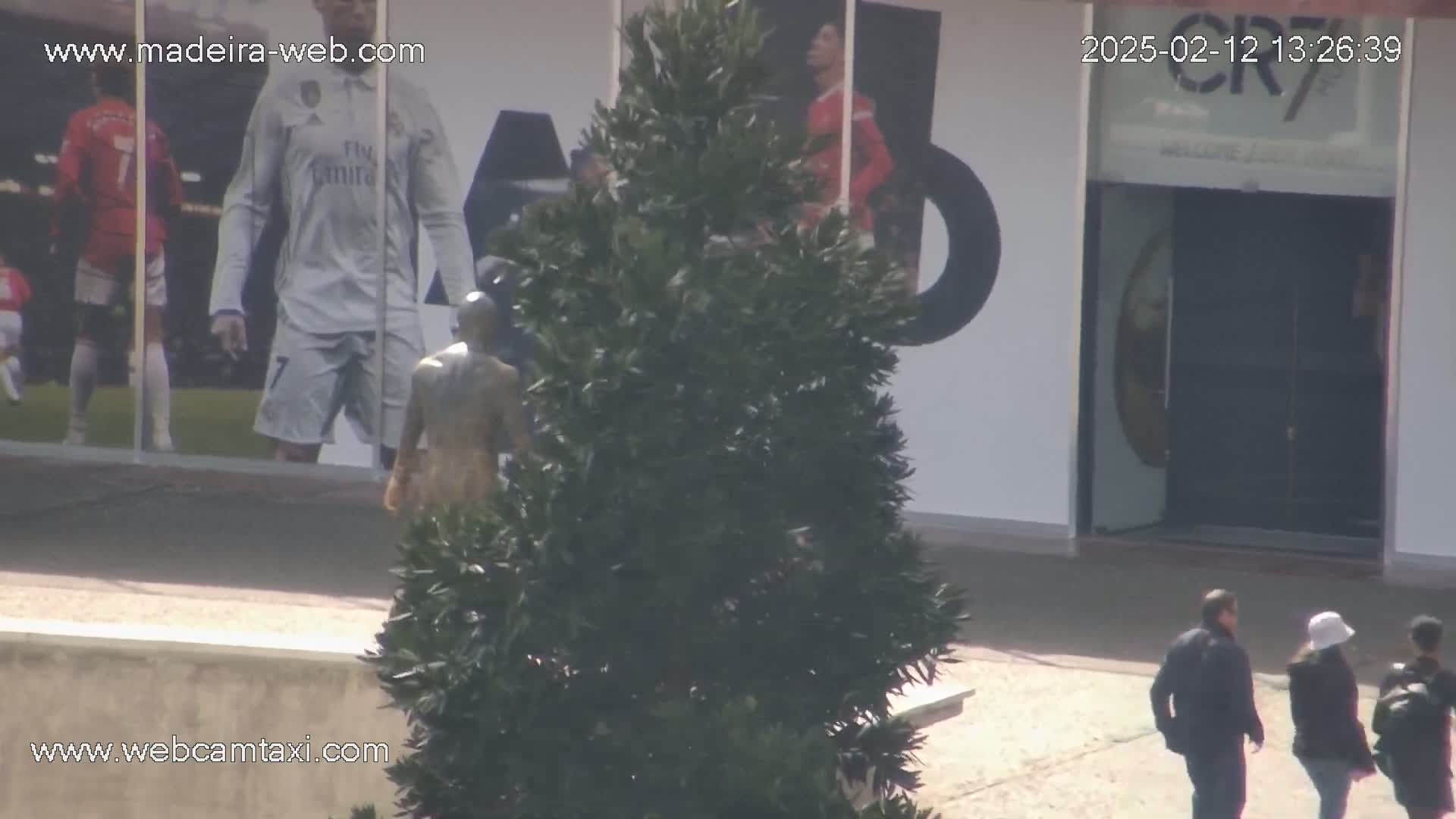A bronze statue stands near a building with large images of soccer players on its exterior, and several people are walking in the sunny outdoor area.