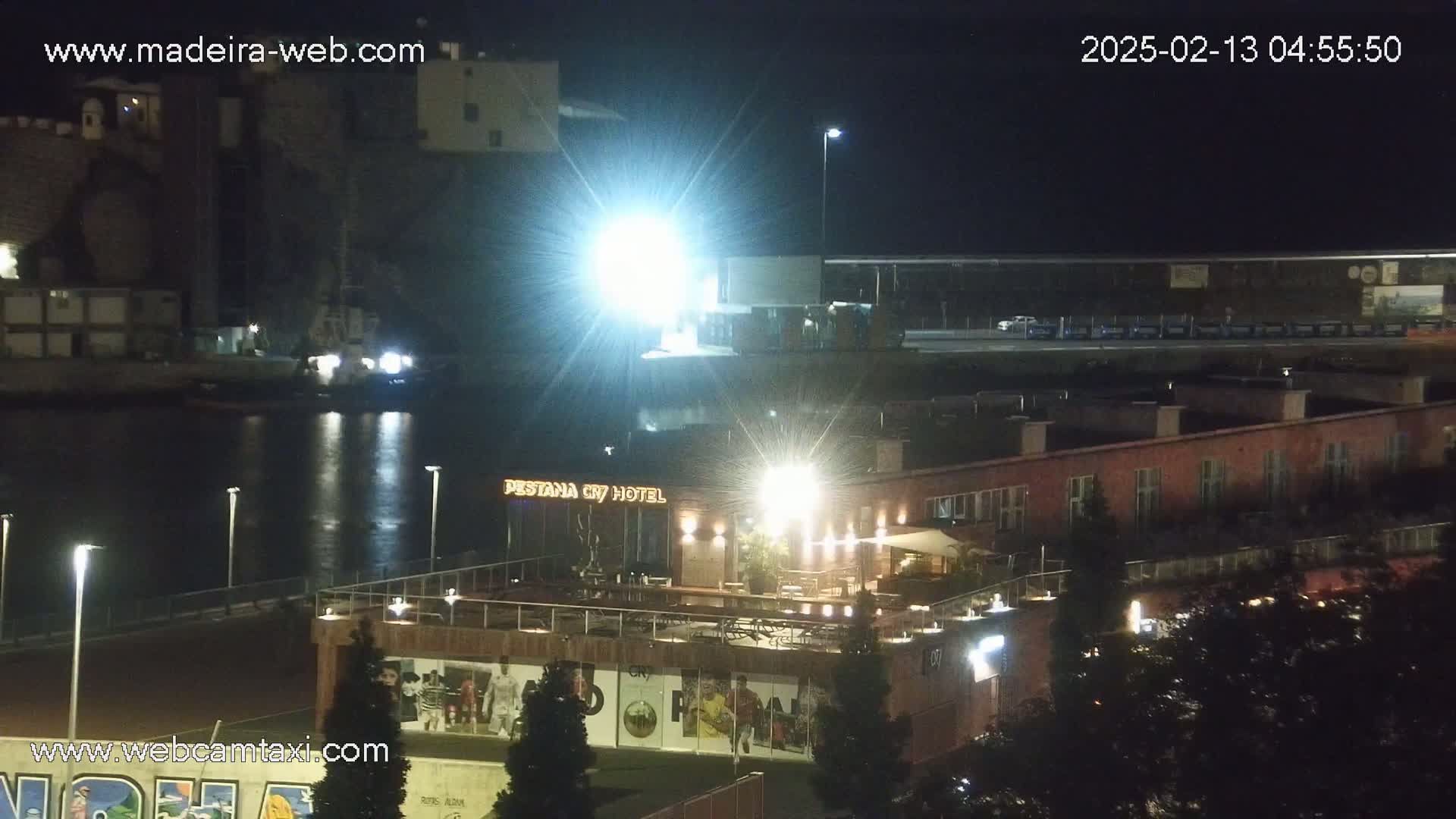 A nighttime view of a waterfront area with a hotel, harbor, and buildings illuminated by bright lights, under clear skies.