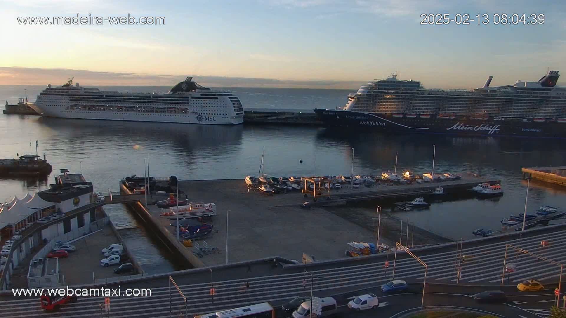 Two large cruise ships are docked at a harbor at sunrise, with several smaller boats moored nearby and cars driving on a street in the foreground.