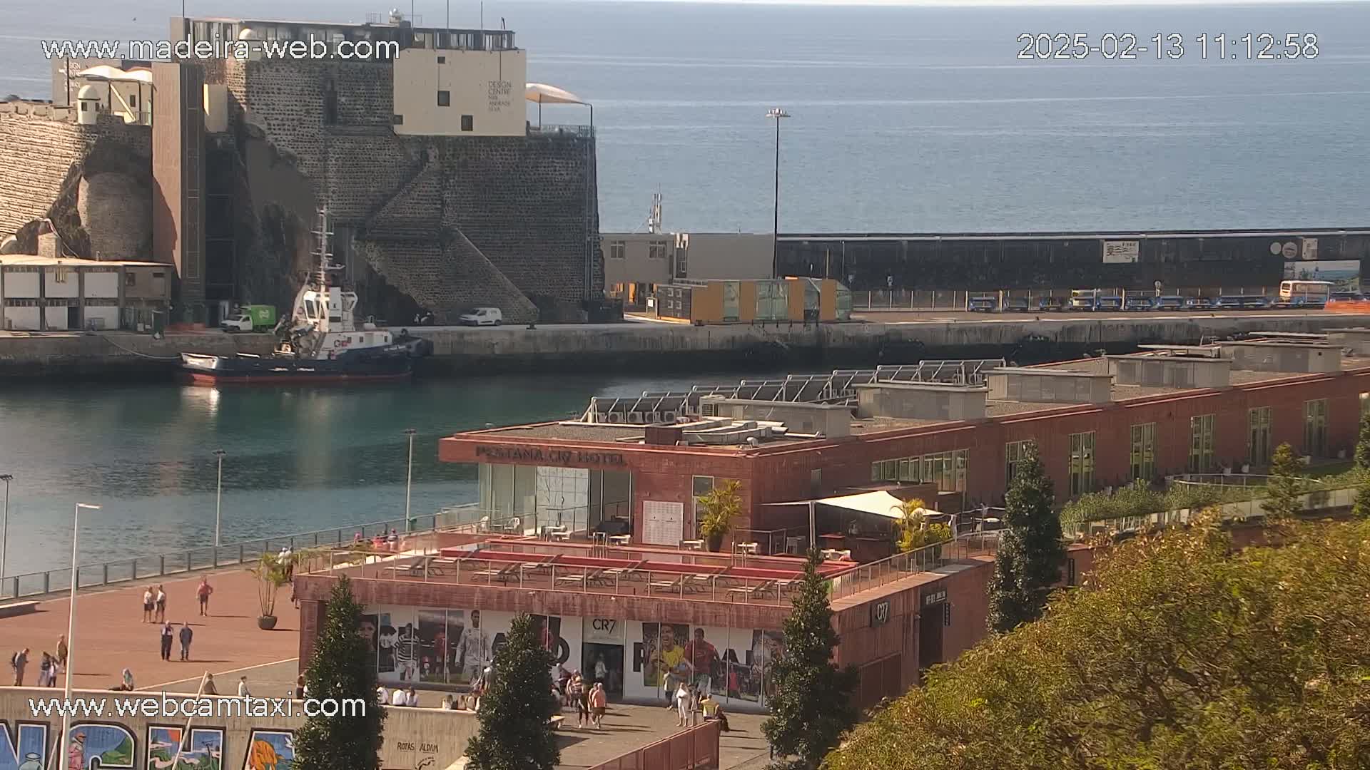 A harbor scene on a sunny day shows a tugboat in the water, a red building with a rooftop terrace, and a calm ocean in the background.