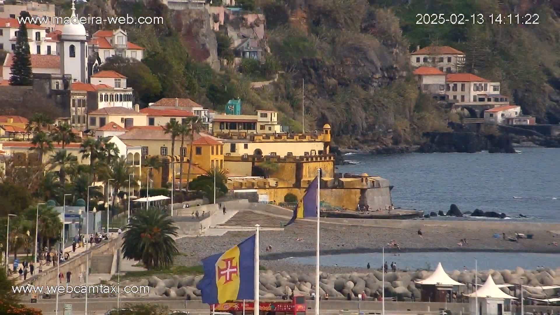 A coastal town with a yellow fortress, a pebble beach, and calm water is seen on a sunny day.