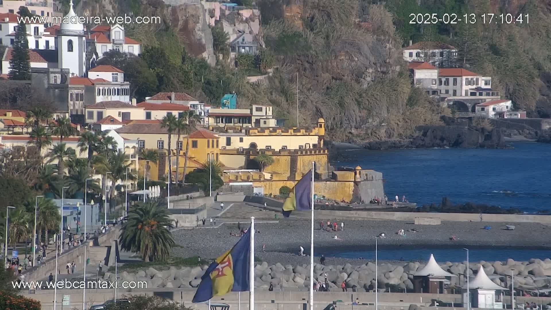 A coastal town with a yellow fortress sits on a rocky hillside overlooking a calm ocean on a sunny day.