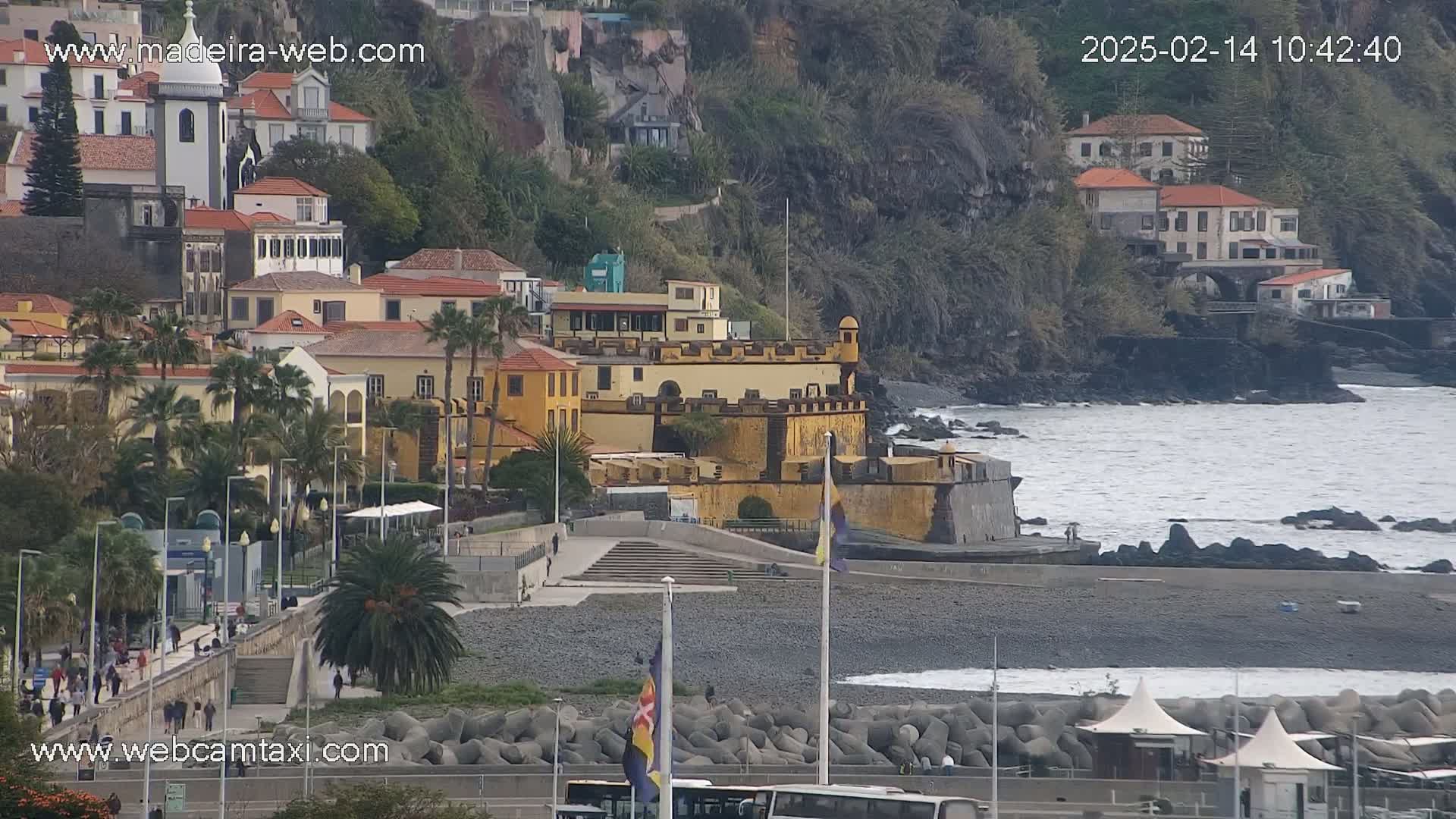 A coastal town with a yellow fortress overlooks a dark-grey pebble beach and a choppy ocean under overcast skies.