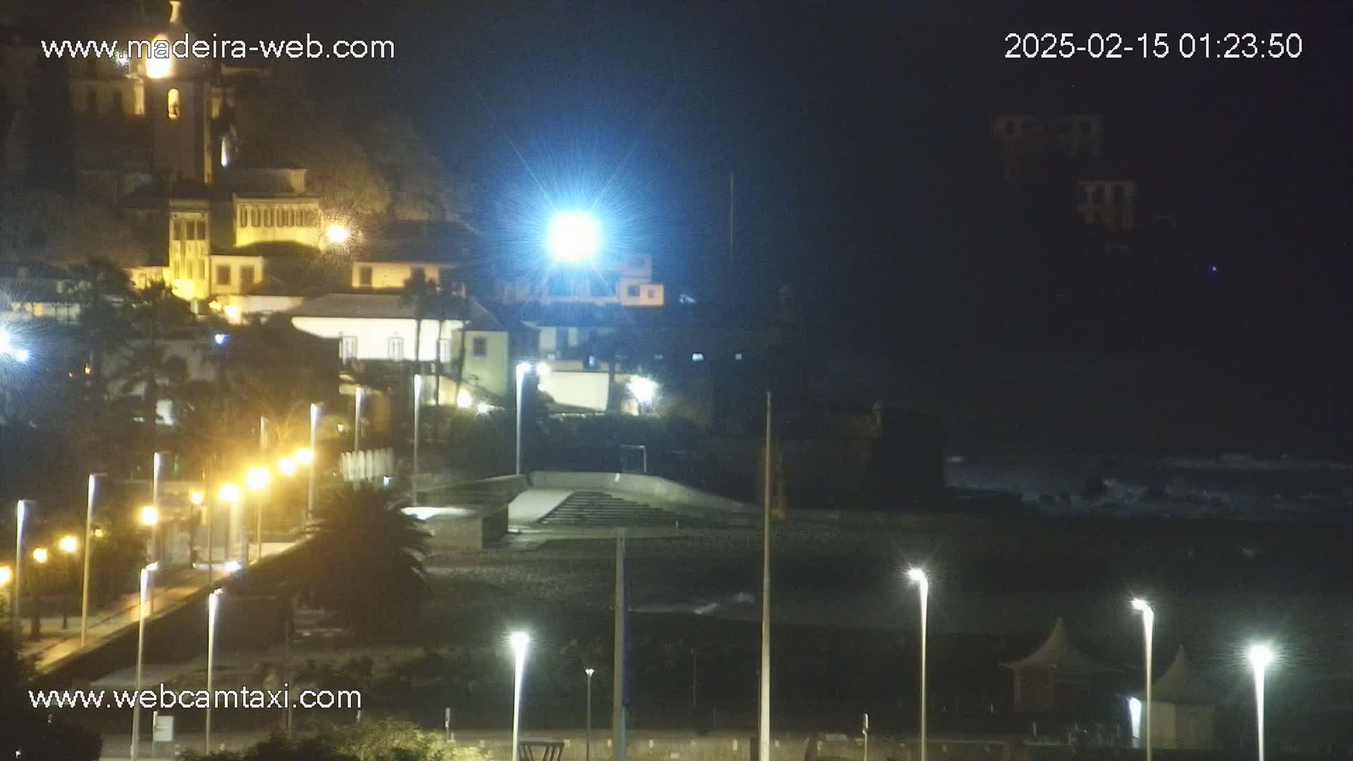 A nighttime view of a coastal town, illuminated by streetlights, with dark, wavy water visible in the distance.