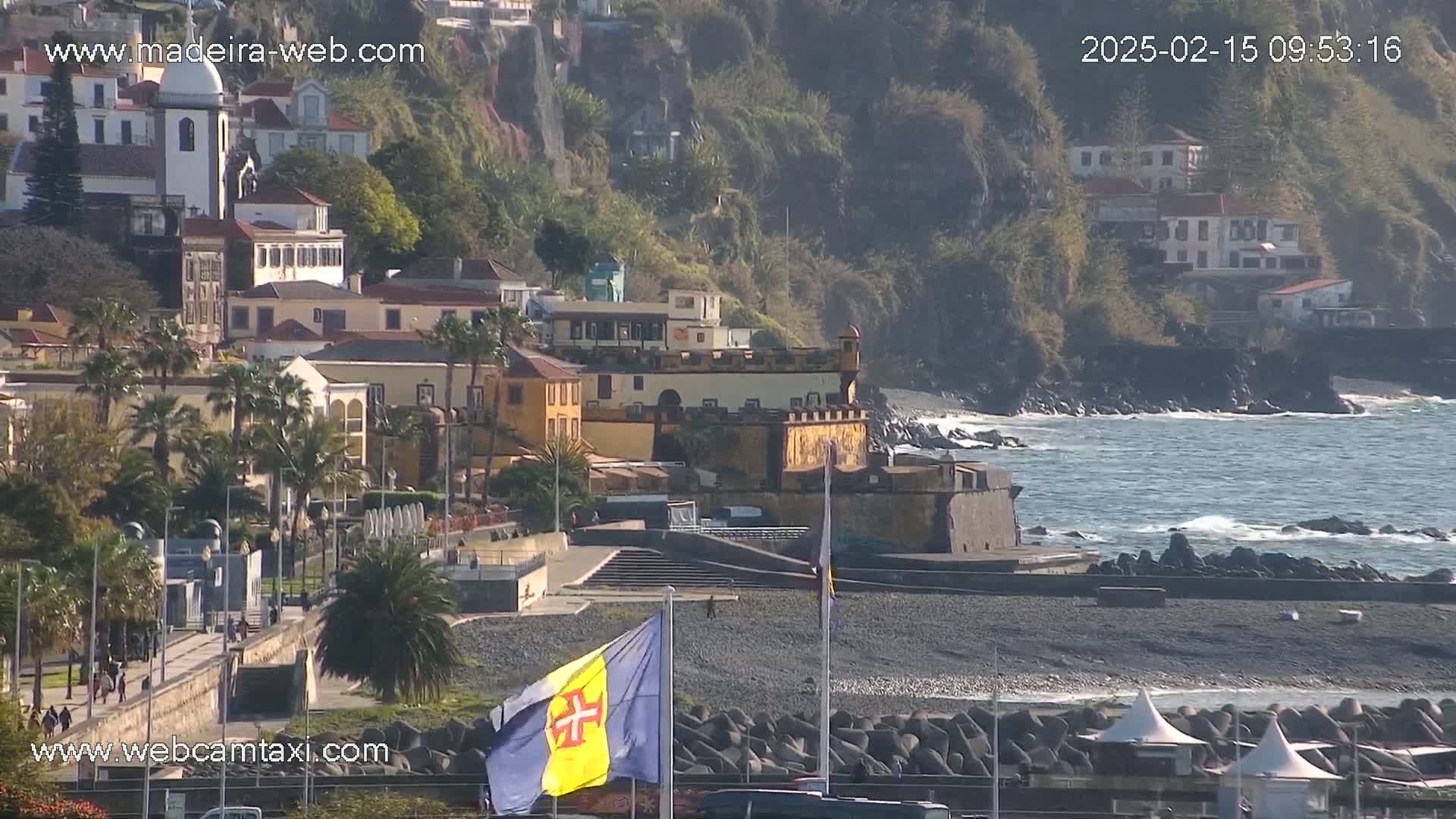 A coastal town with a fortress and a flag sits on a dark-sand beach next to a rocky shoreline and a somewhat choppy ocean under a sunny sky.