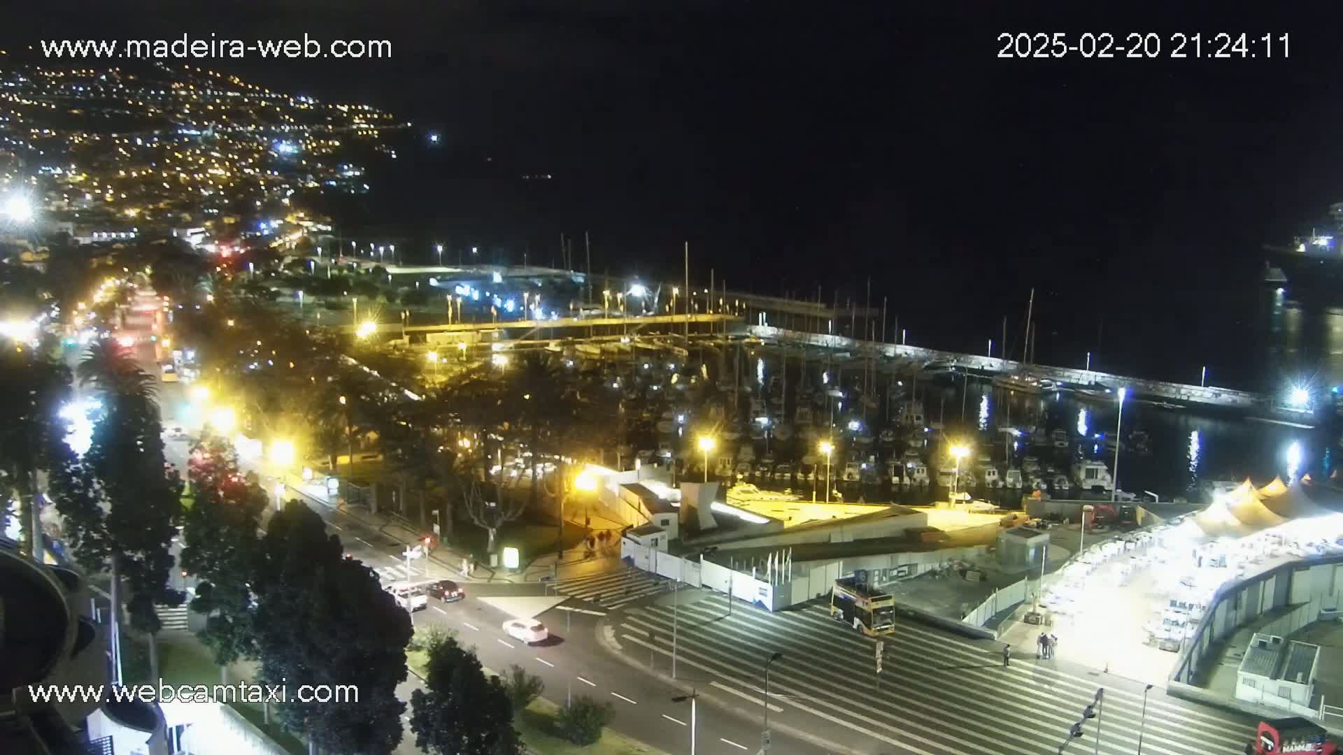 A nighttime aerial view of a harbor filled with boats, surrounded by city lights and roads with cars.