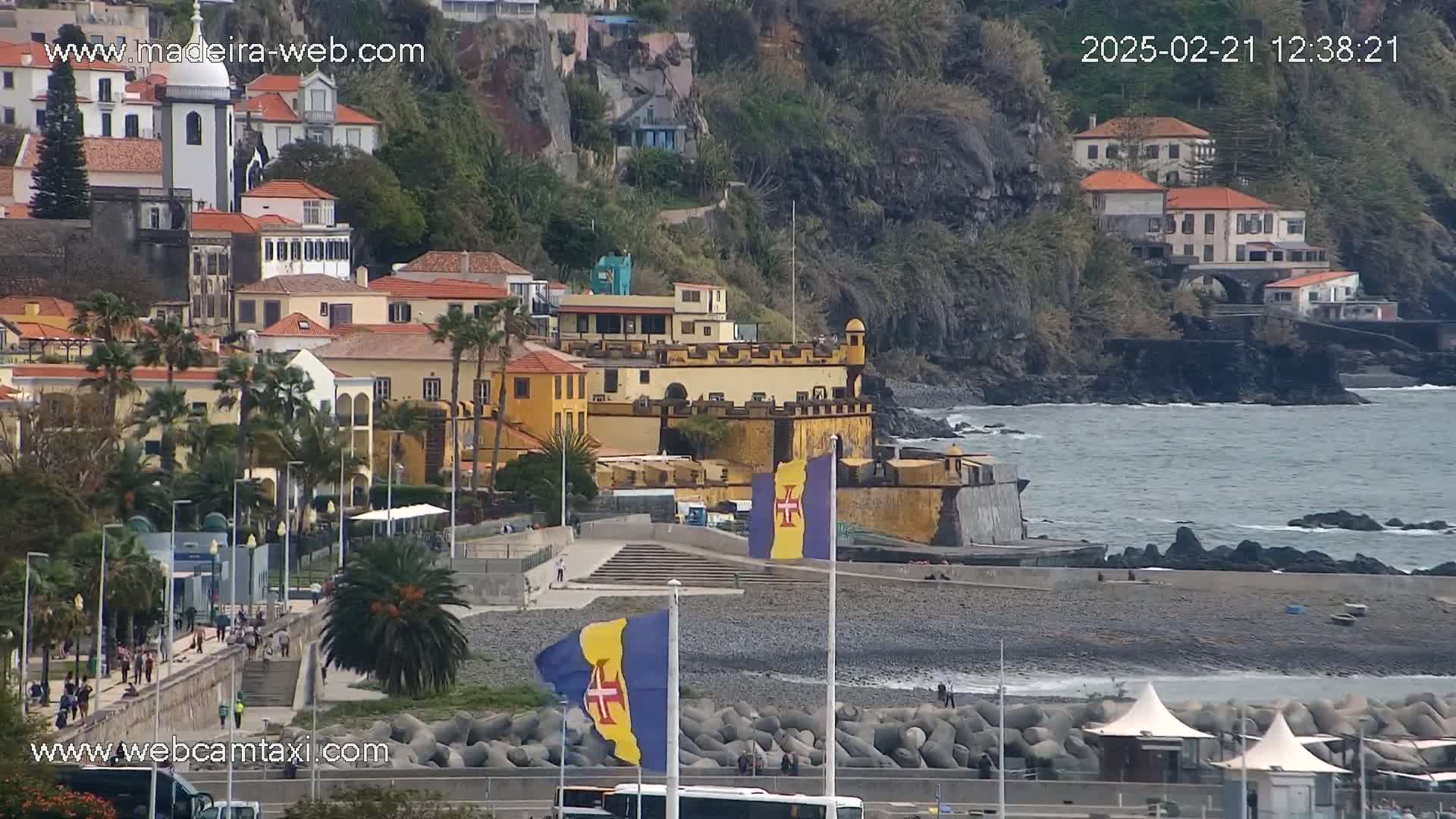 A coastal town with a yellow fort sits on a rocky shore next to a calm ocean under an overcast sky.