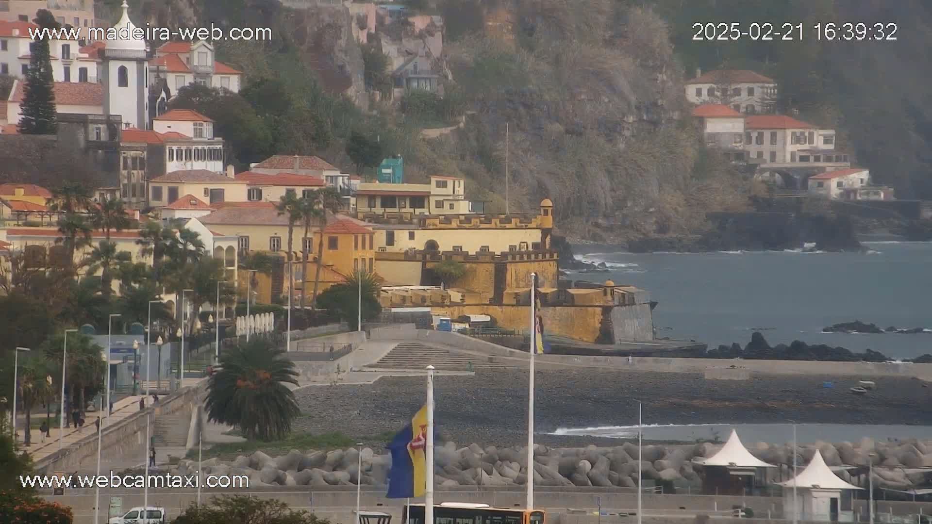 A coastal town with colorful buildings, a fortress, and a rocky beach is seen on an overcast day.